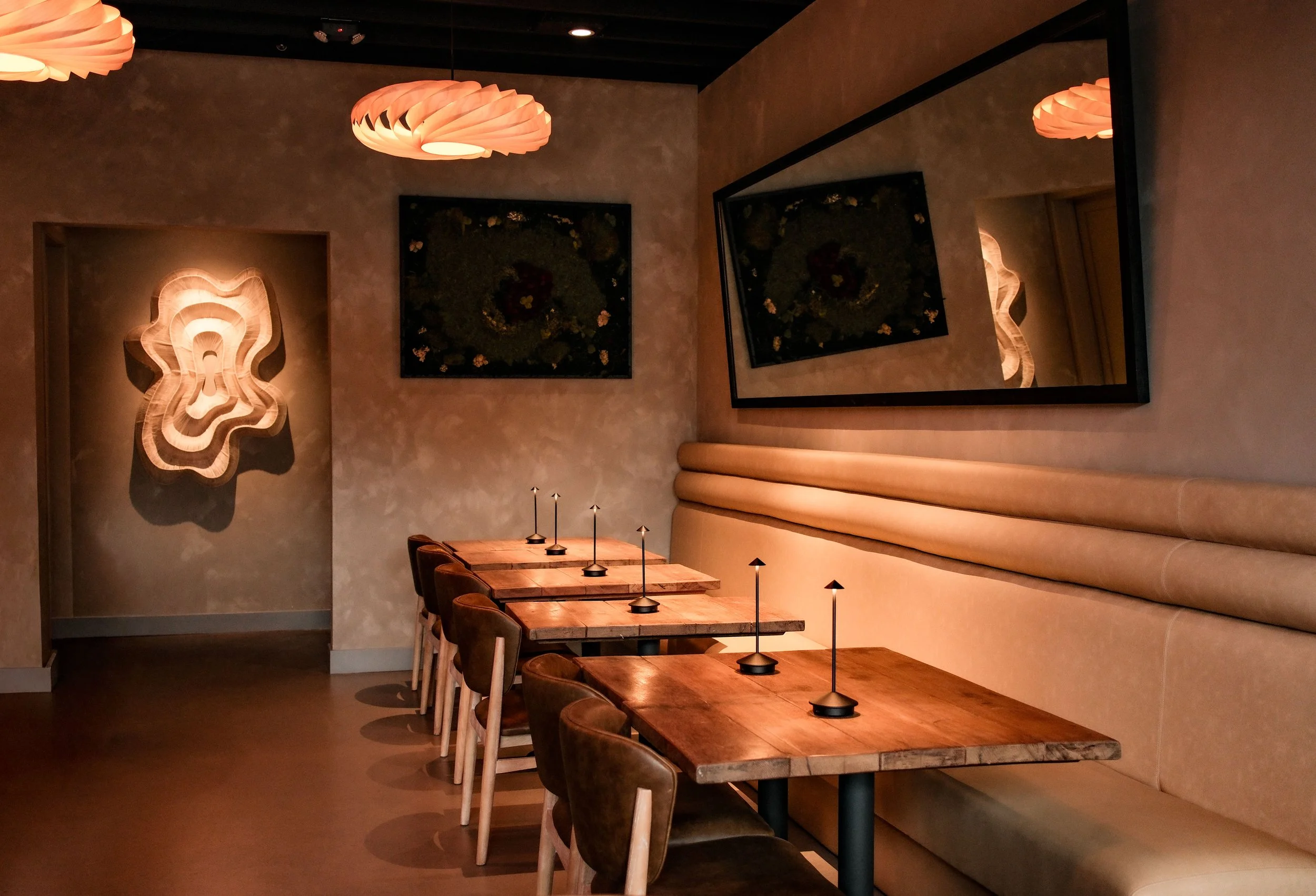Interior of a cozy restaurant with wooden tables, leather chairs, wall art, a large mirror, and warm lighting fixtures.