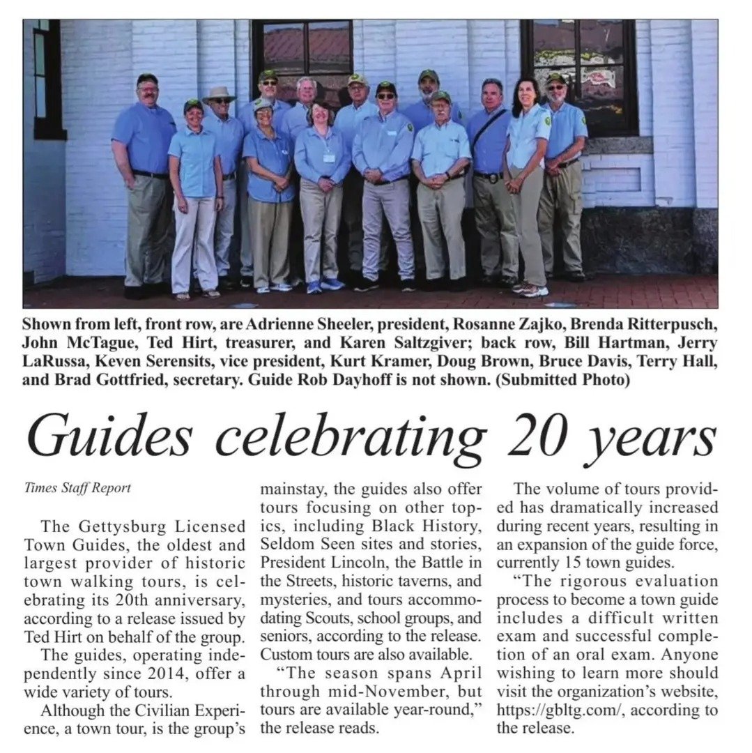 Photograph of a group of fifteen Tour guides standing outdoors in front of a white building with windows, posing for a group photo. The group includes both men and women, dressed in casual attire with some wearing hats and sunglasses.