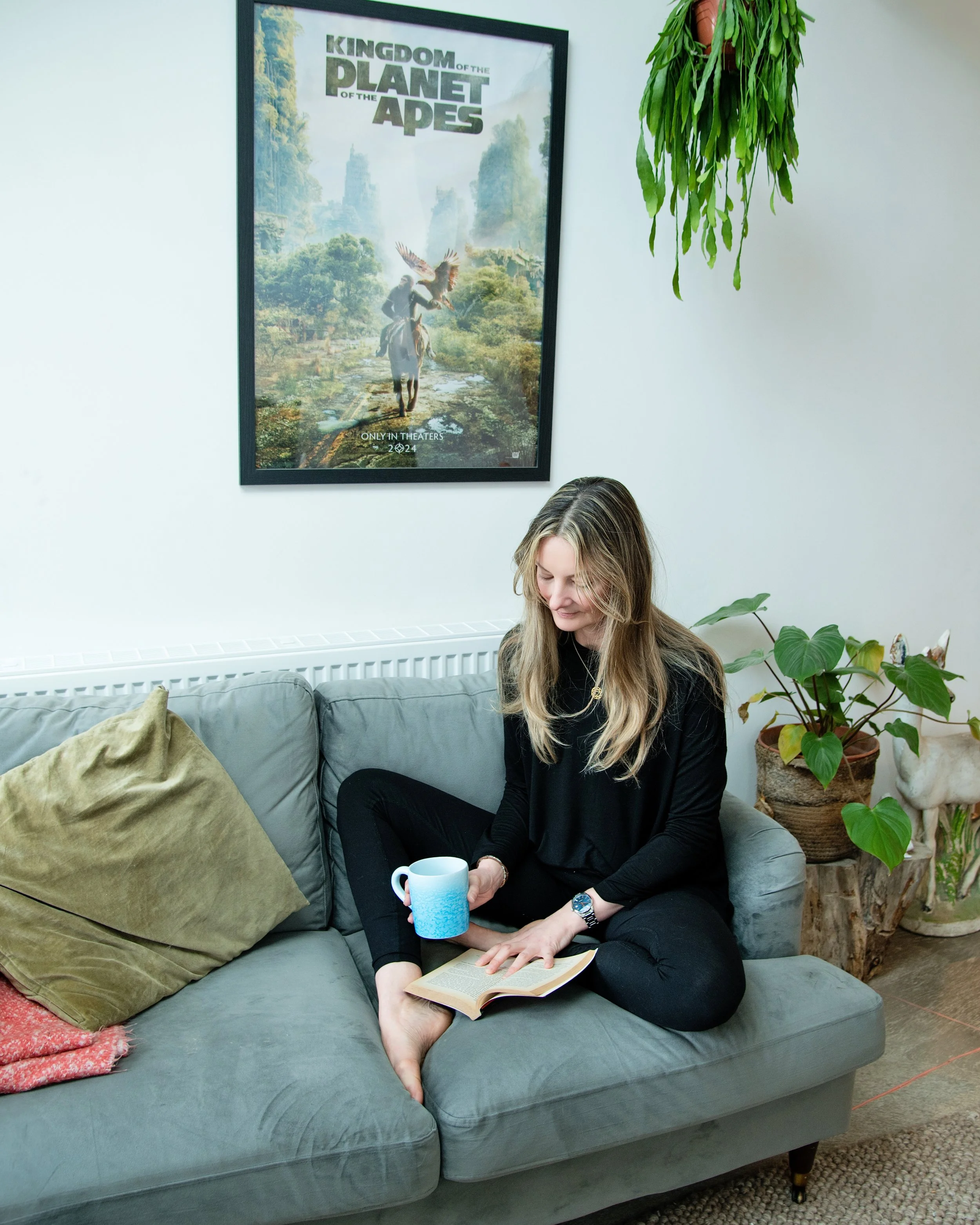A calm yoga woman sitting on a grey couch in a living room, holding a blue mug and reading a book, with plants nearby and a 'Kingdom of the Planet of the Apes' movie poster on the wall.