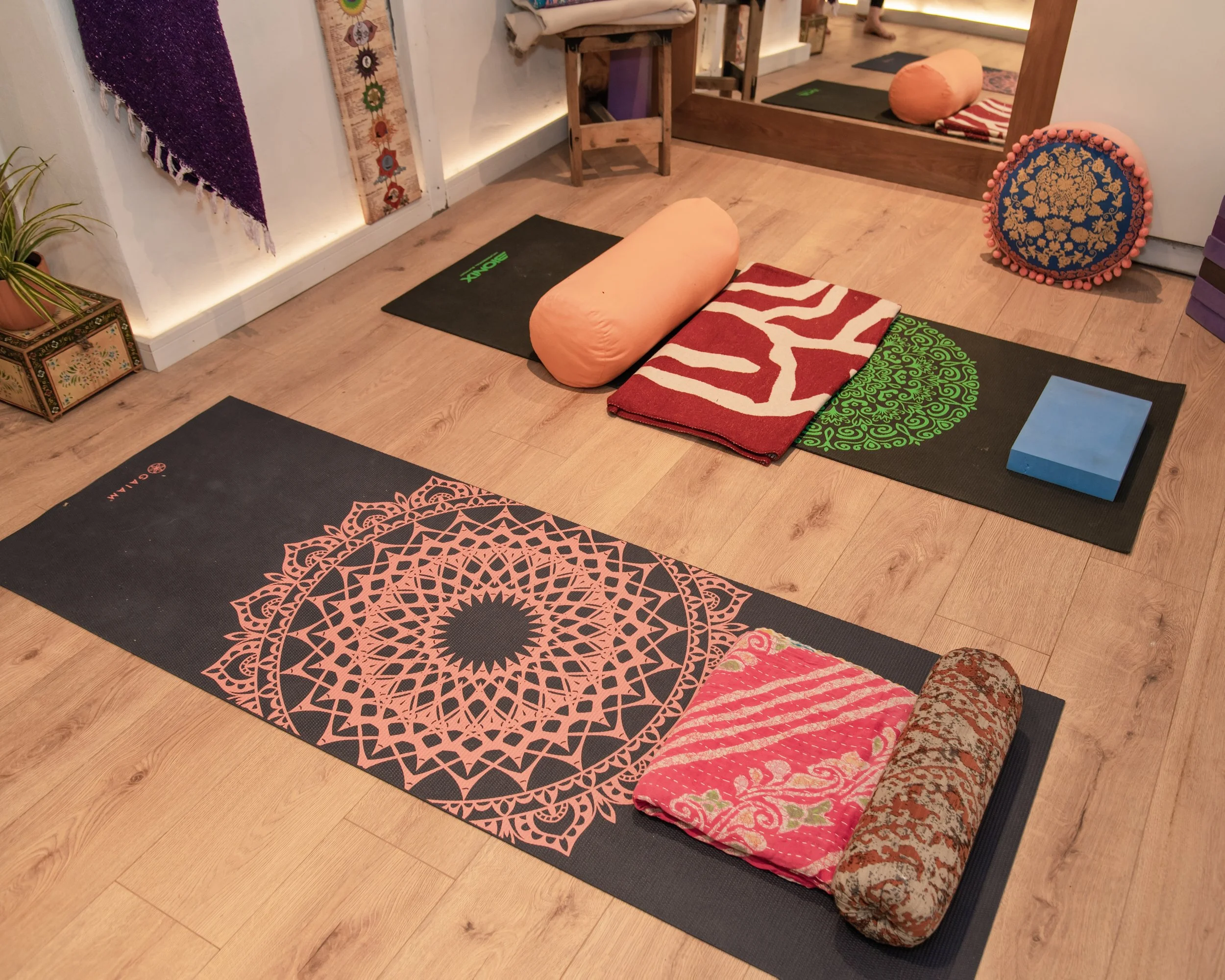 Yoga room with two mats, blankets, bolster pillow, blocks, and decorative cushions, with a mirror on the wall and plants in the corner.