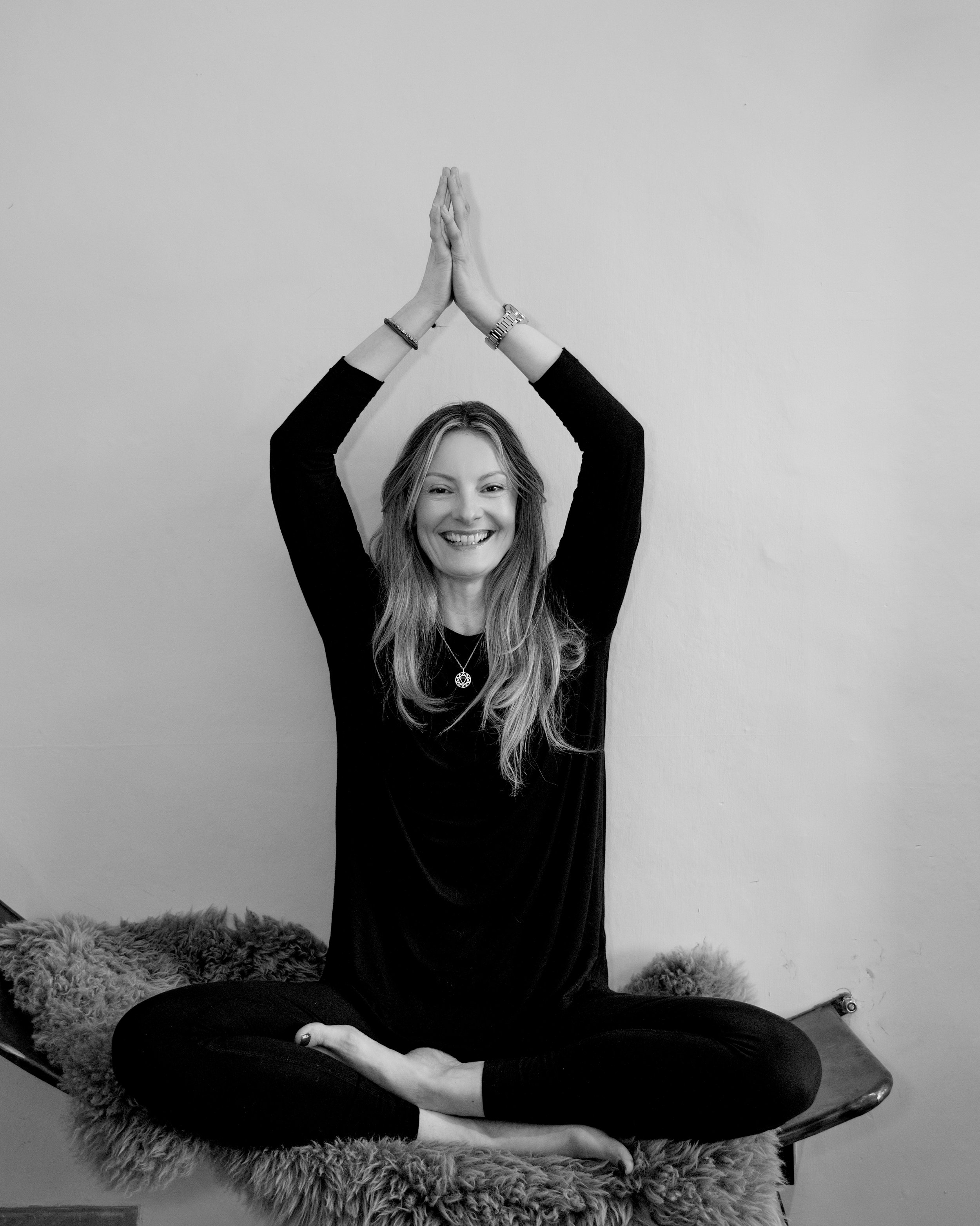 A yoga instructor sitting cross-legged on a furry cushion, smiling, with her arms raised above her head and palms pressed together in a yoga pose, against a plain wall.