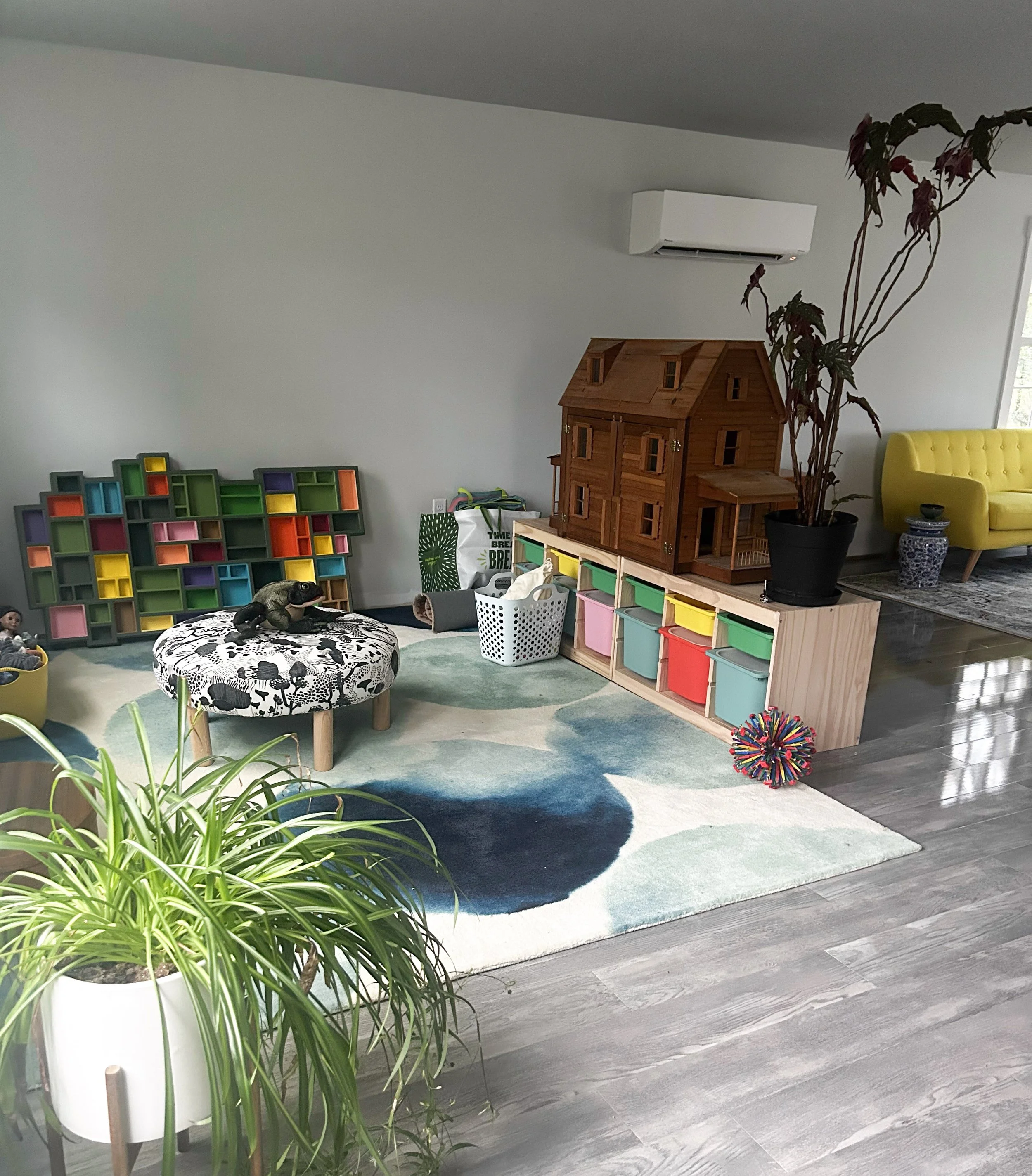 Play Therapy room with a colorful bookshelf, storage bins, a dollhouse, and a yellow couch, decorated with plants and a patterned rug.