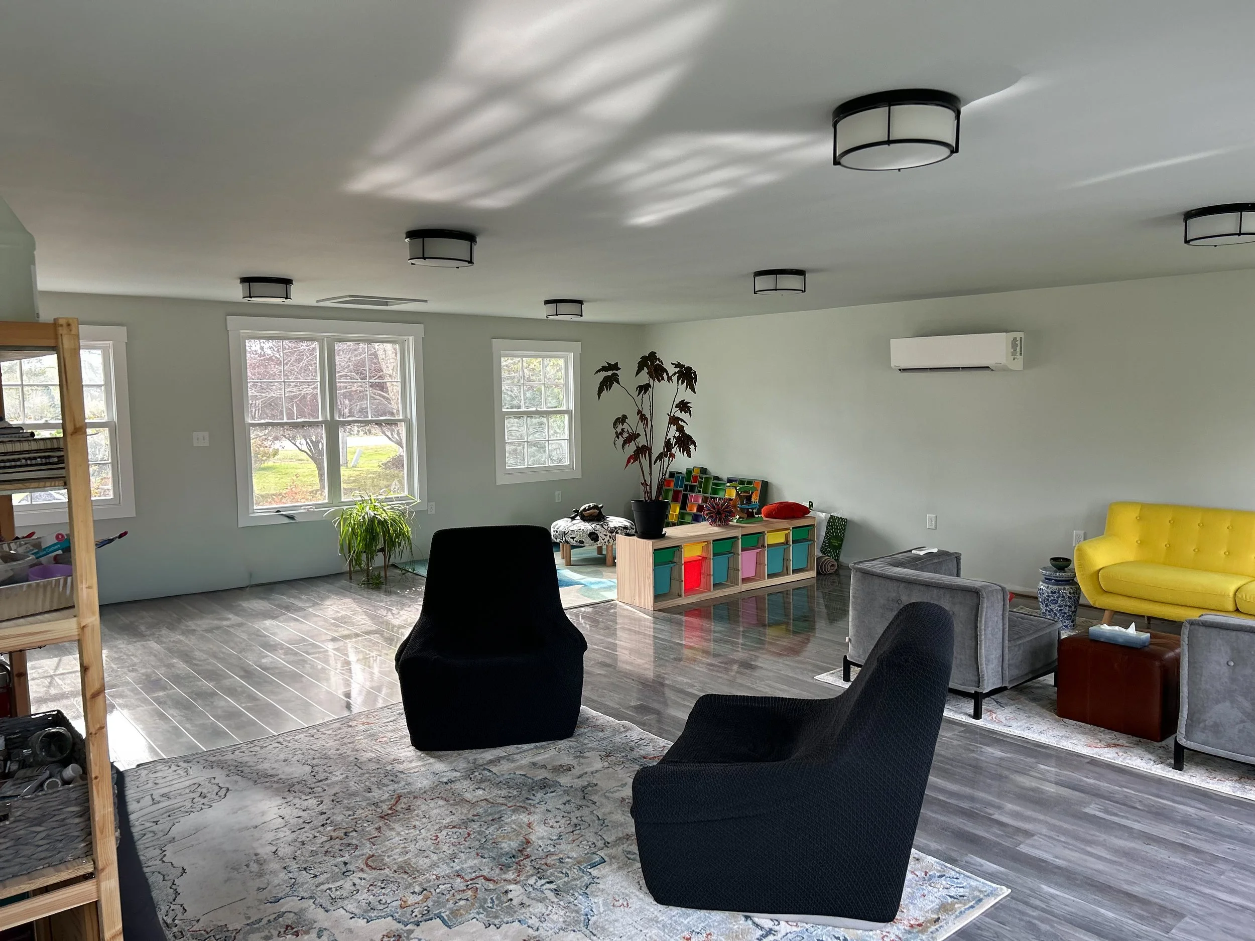 Art Therapy room with large windows, gray flooring, black and gray chairs, a yellow sofa, a colorful toy storage, and an air conditioning unit on the wall.