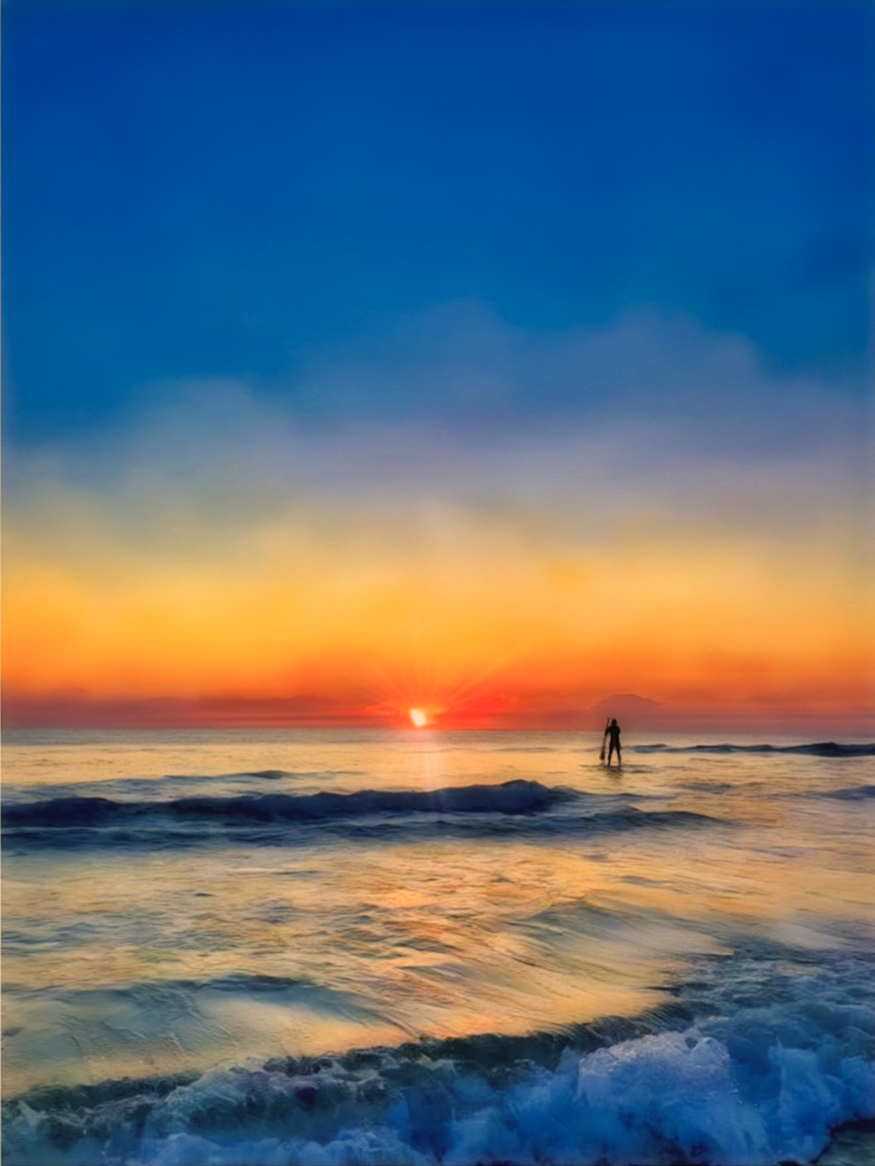 Paddle boarding  in the ocean, against the early morning sunrise, with a colorful sky including orange, yellow, red, and blue, hues.