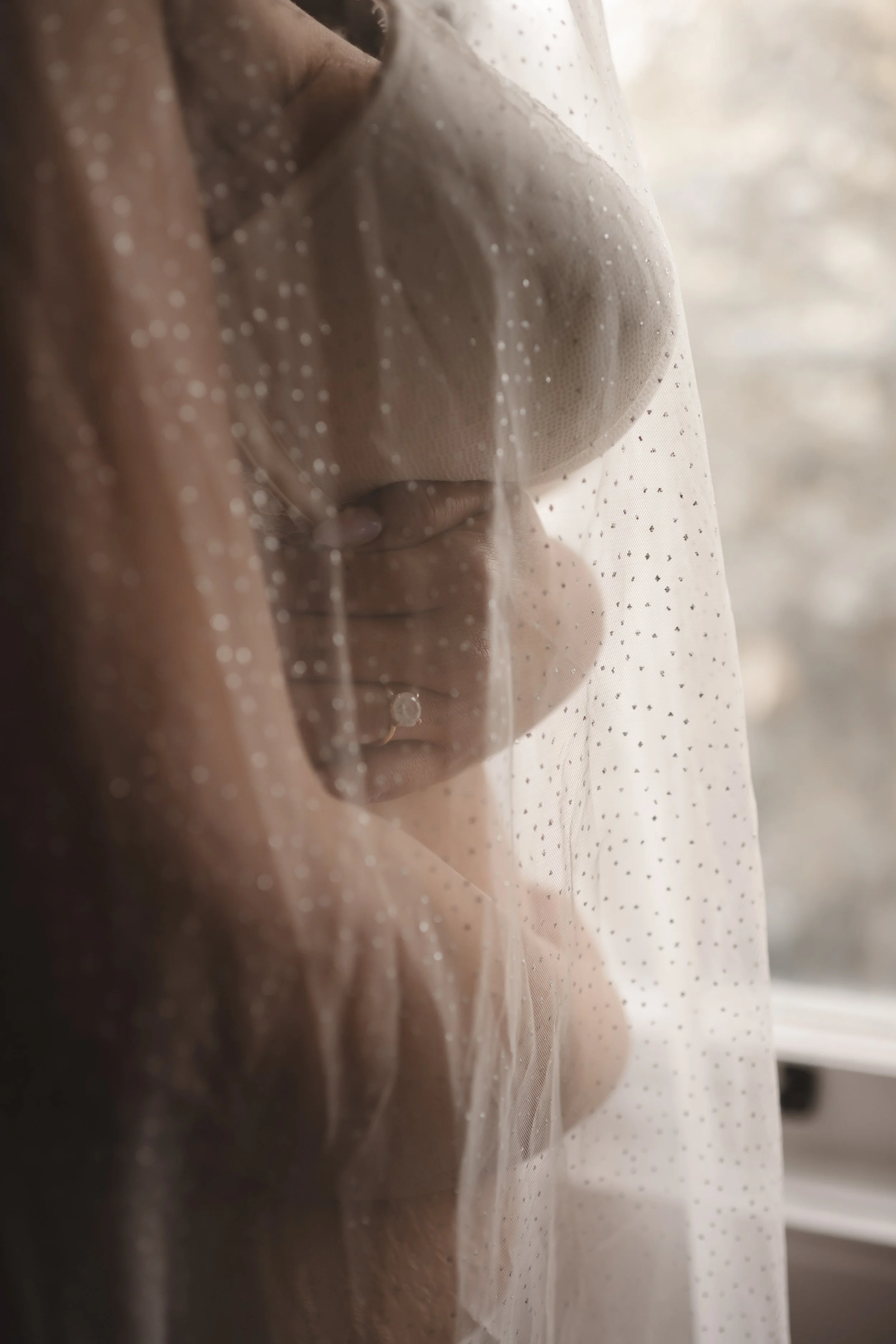 A woman holding her hand with a ring over her chest behind a sheer veil with small dots, near a window with bright light.