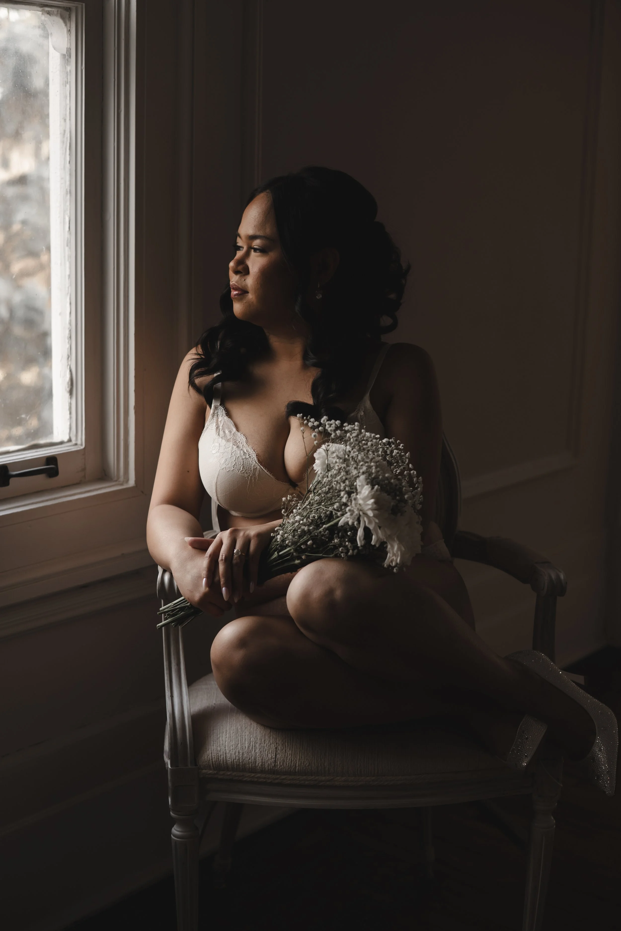 A woman holding a bouquet of flowers sitting on a vintage-style chair by a window, wearing lingerie, with soft lighting and a contemplative expression.