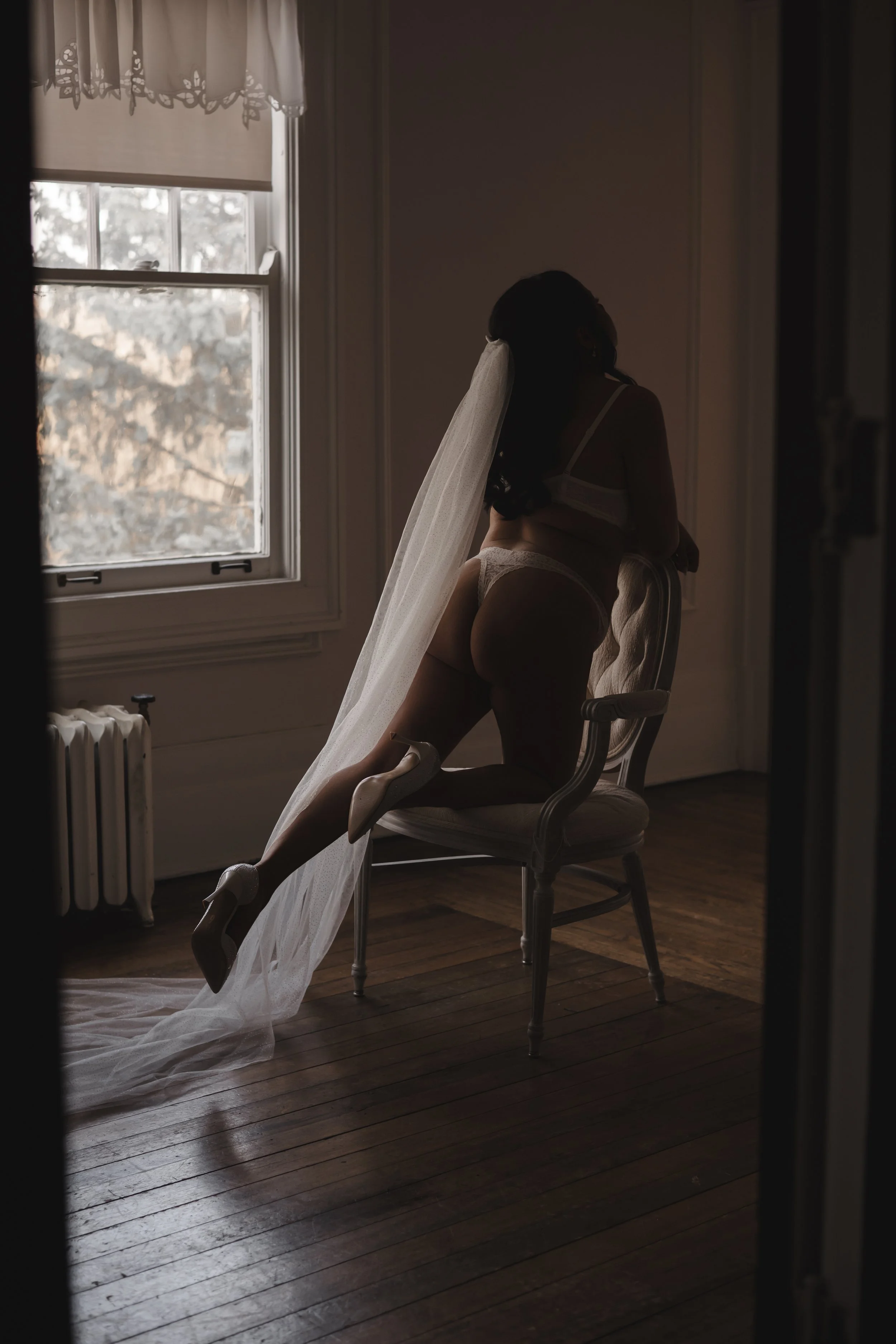 A woman in lingerie, a veil, and high heels poses on a chair in a dimly lit room near a window with curtains, with sunlight coming through.