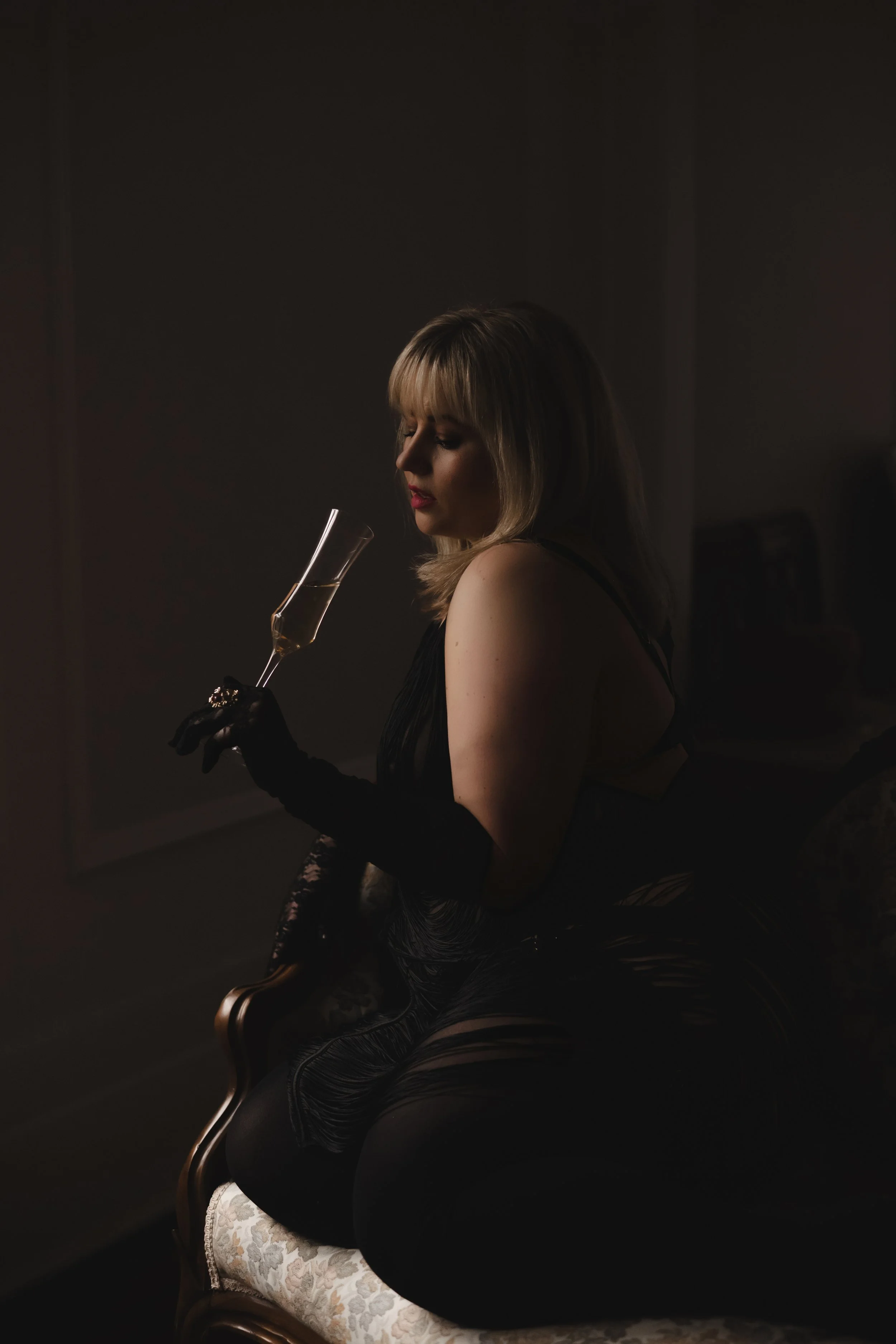 A woman with blonde hair in black lace clothing and long black gloves sitting on a vintage chair, holding a champagne flute, in a dimly lit room.