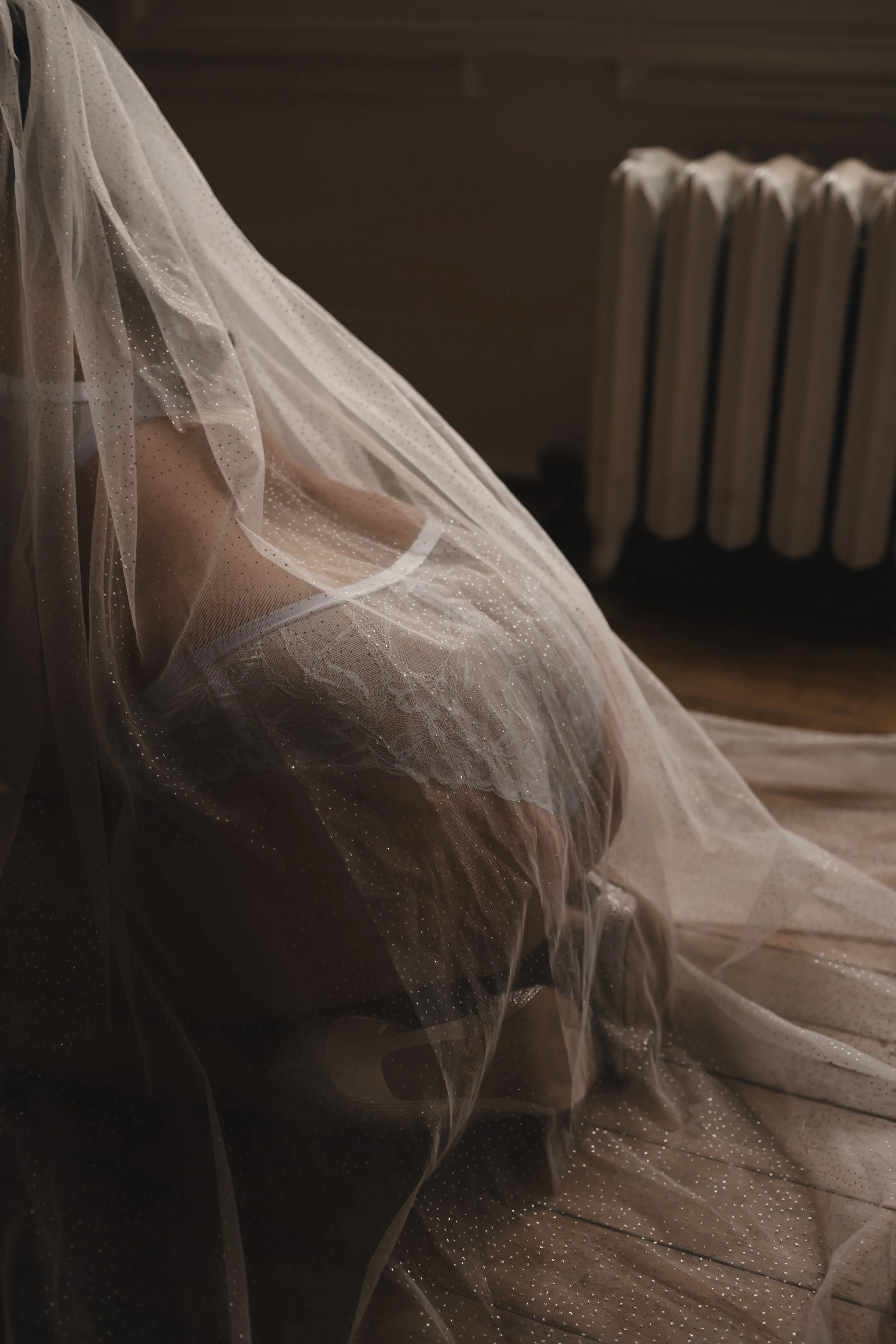 Person kneeling on the wooden floor, covered by a sheer, sparkly veil or fabric, in front of a dark wall and a radiator.