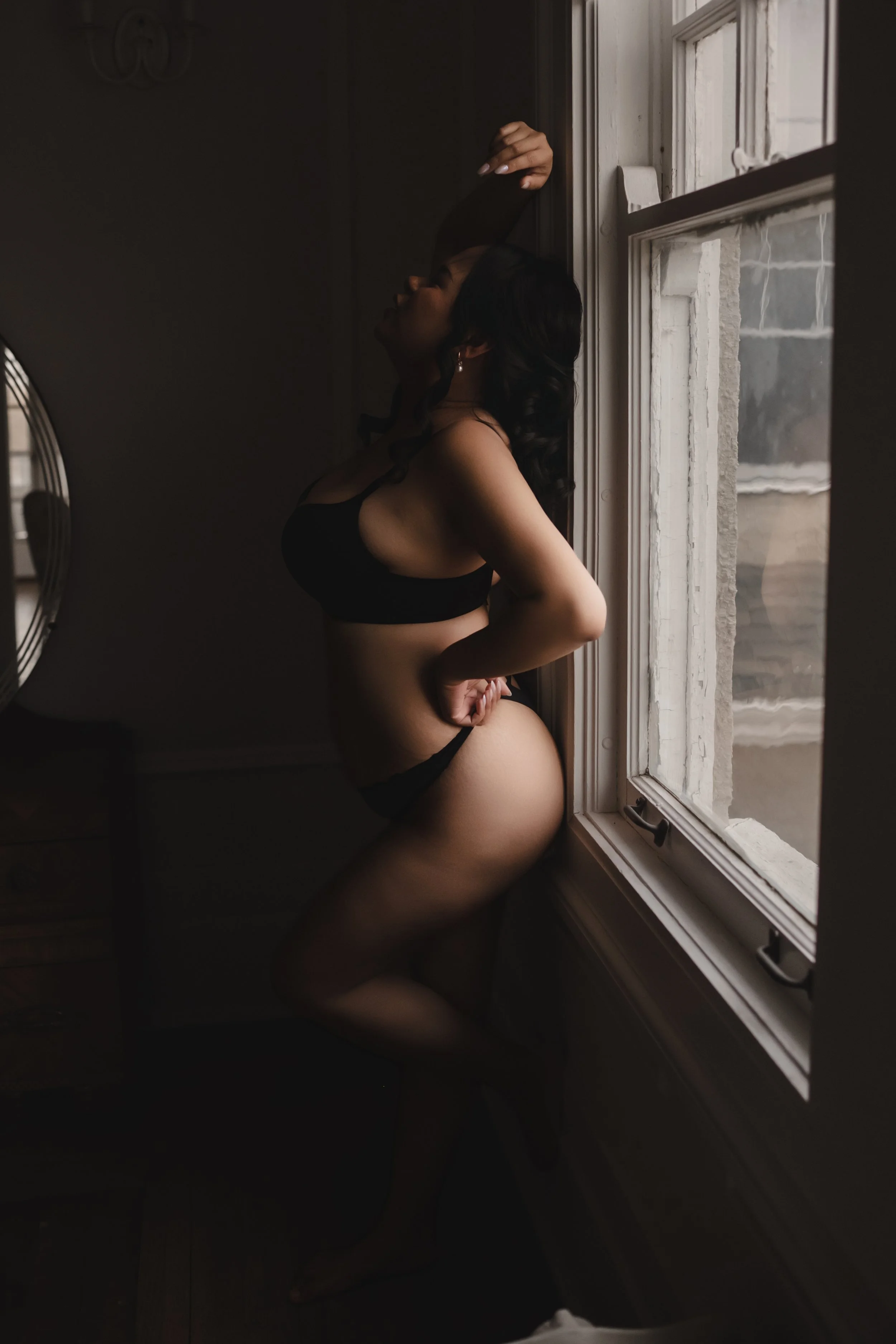 A woman standing by a window in a dimly lit room, wearing black lingerie and touching her waist.