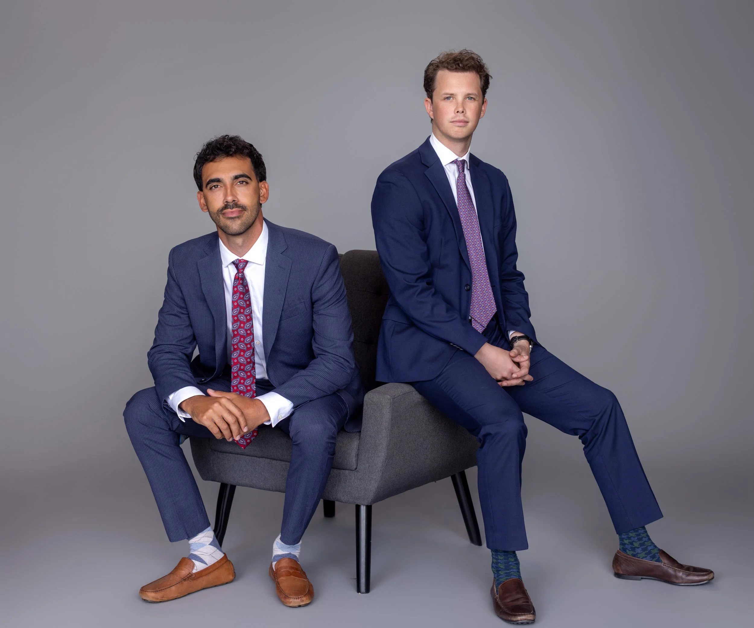 Two men in suits sitting on a small gray sofa against a plain neutral background, with one sitting on the sofa and the other standing beside it.