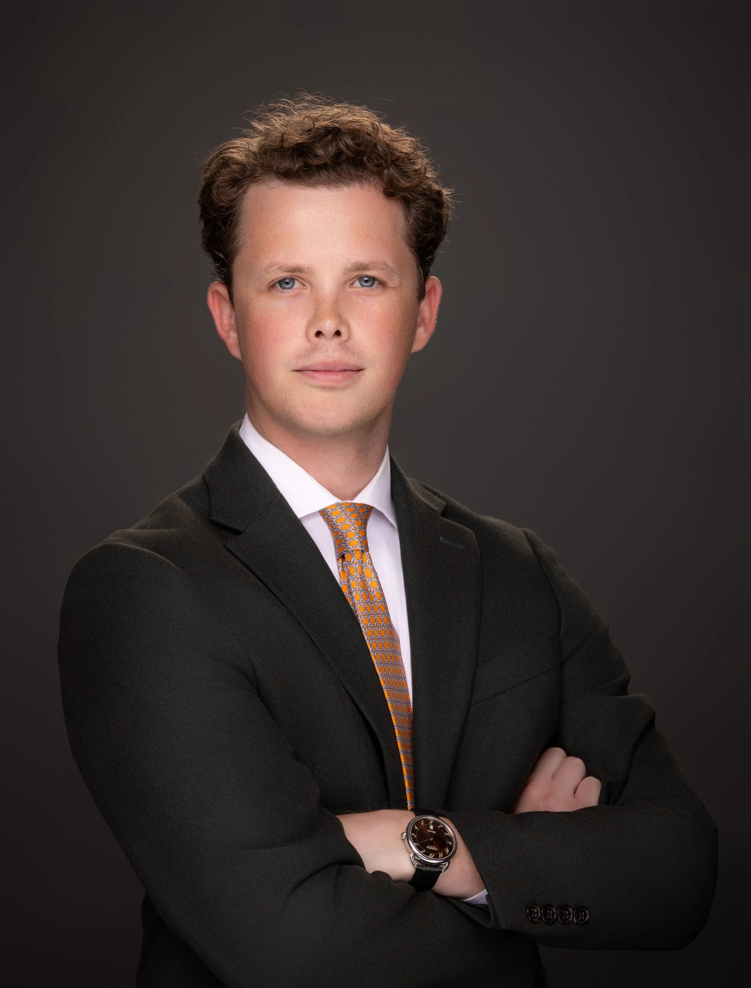 A professional man with curly brown hair, blue eyes, and fair skin posing in front of a dark background. He is wearing a black suit, white shirt, orange patterned tie, and a watch on his left wrist, with his arms crossed.