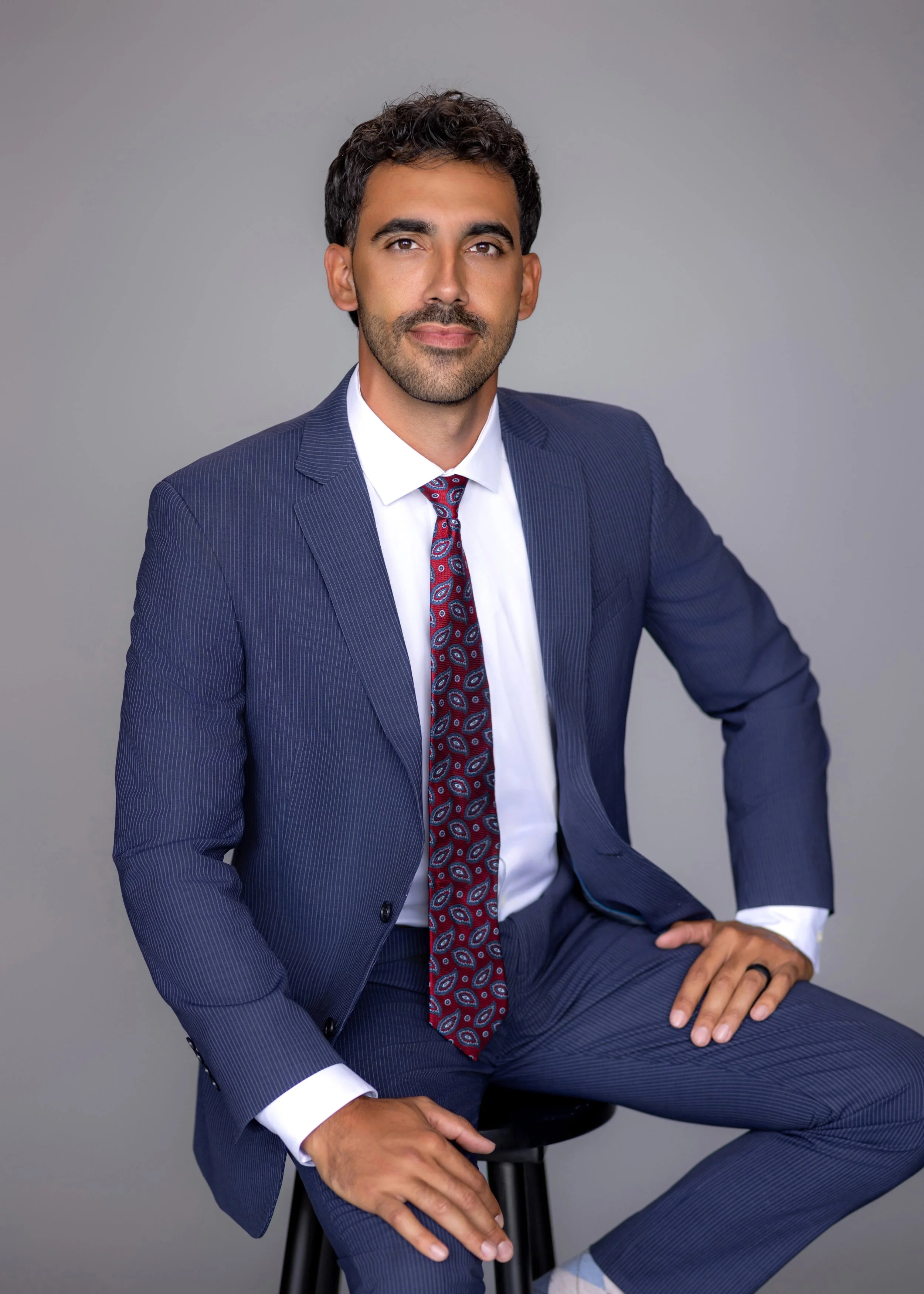 Man in navy pinstripe suit with white shirt and patterned tie sitting on a stool against a gray background.