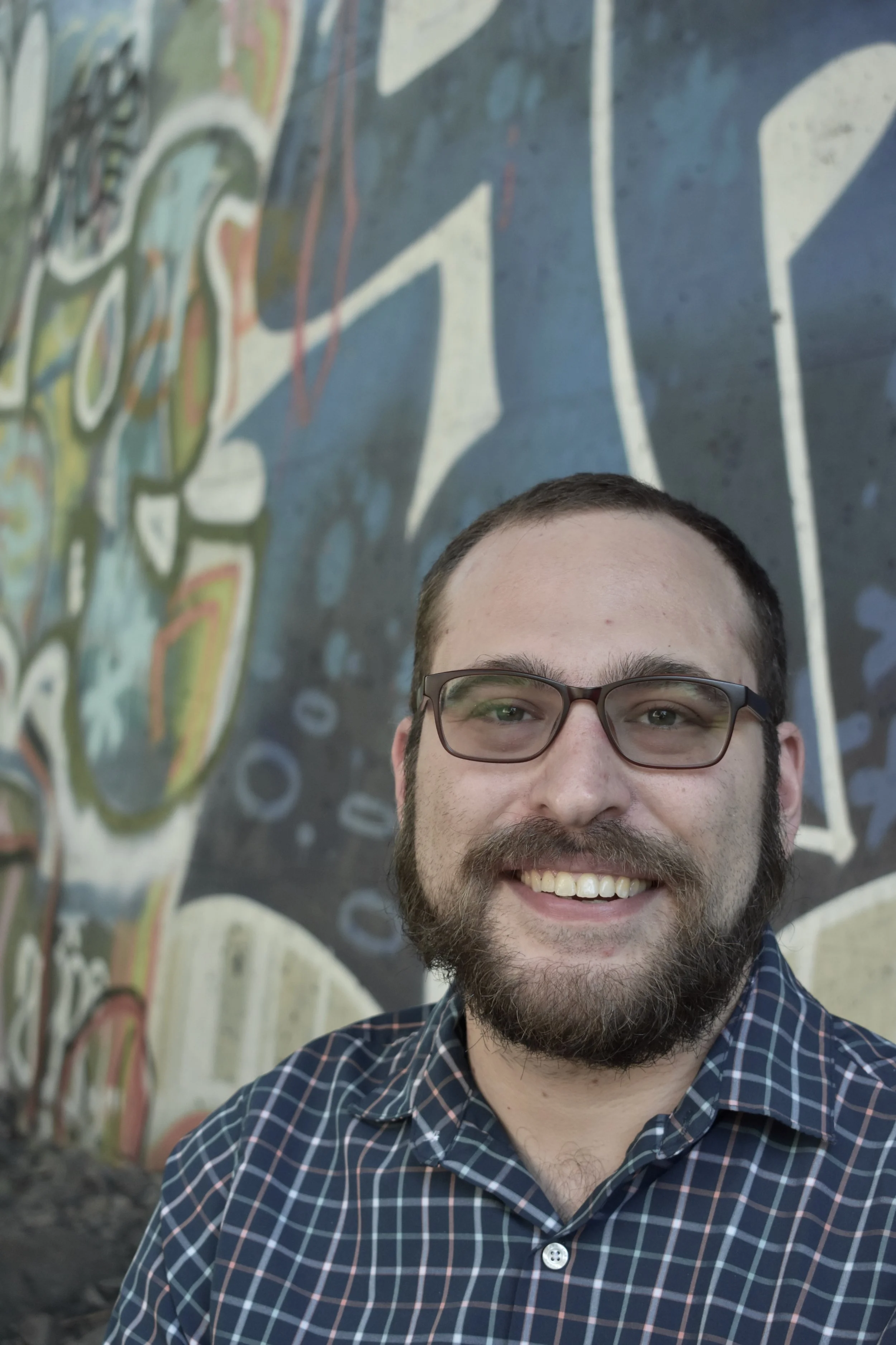 A bearded white man with glasses in a blue checkered shirt with graffiti behind him