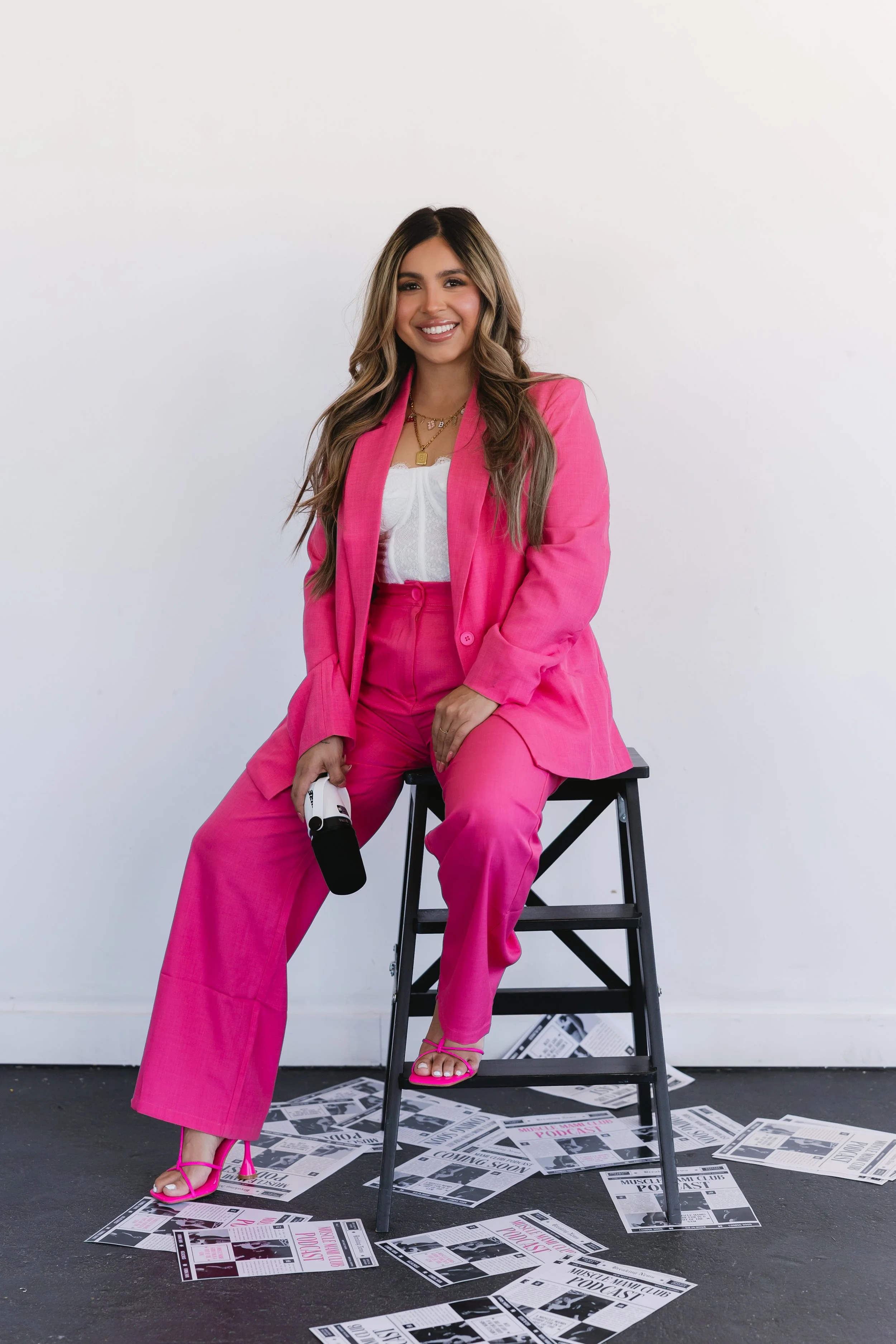 Woman in a pink suit seated on a stool, holding a microphone, surrounded by scattered papers.