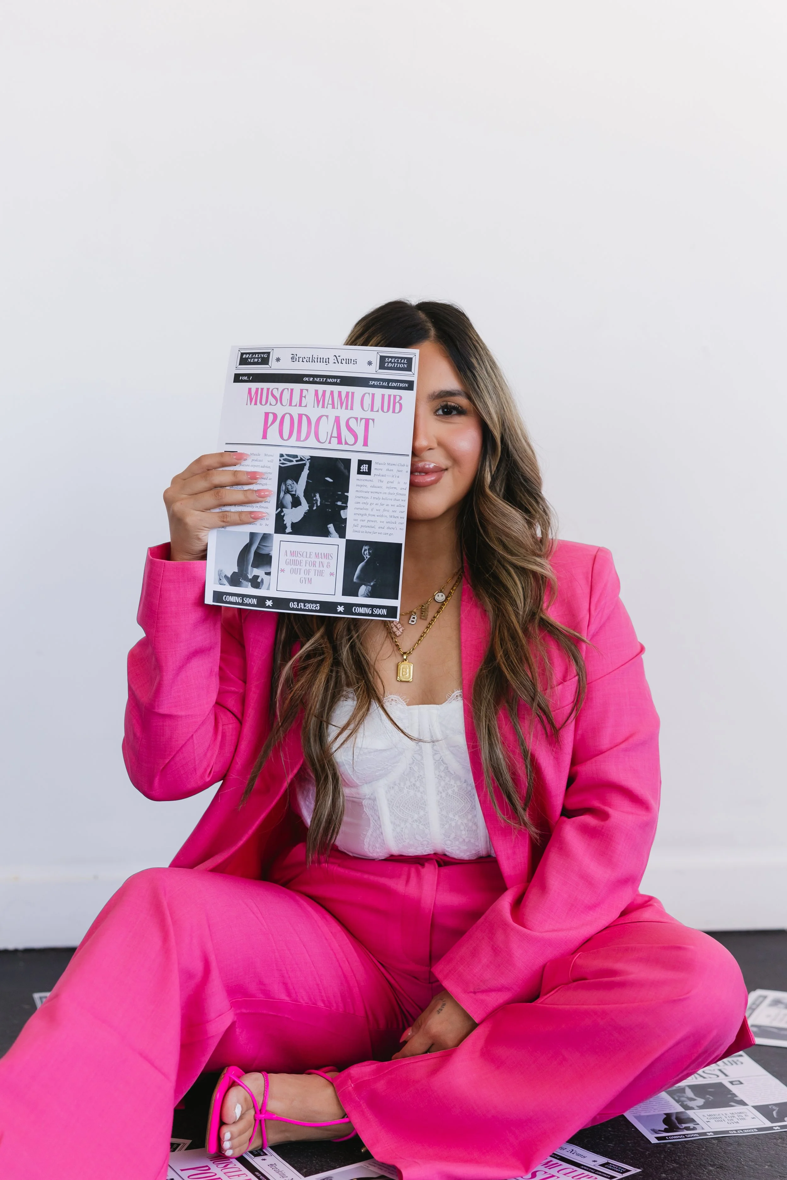 Woman in a pink suit sitting on the floor holding a paper promoting "Muscle Mami Club Podcast." The paper covers part of her face, and various papers lie scattered around her.