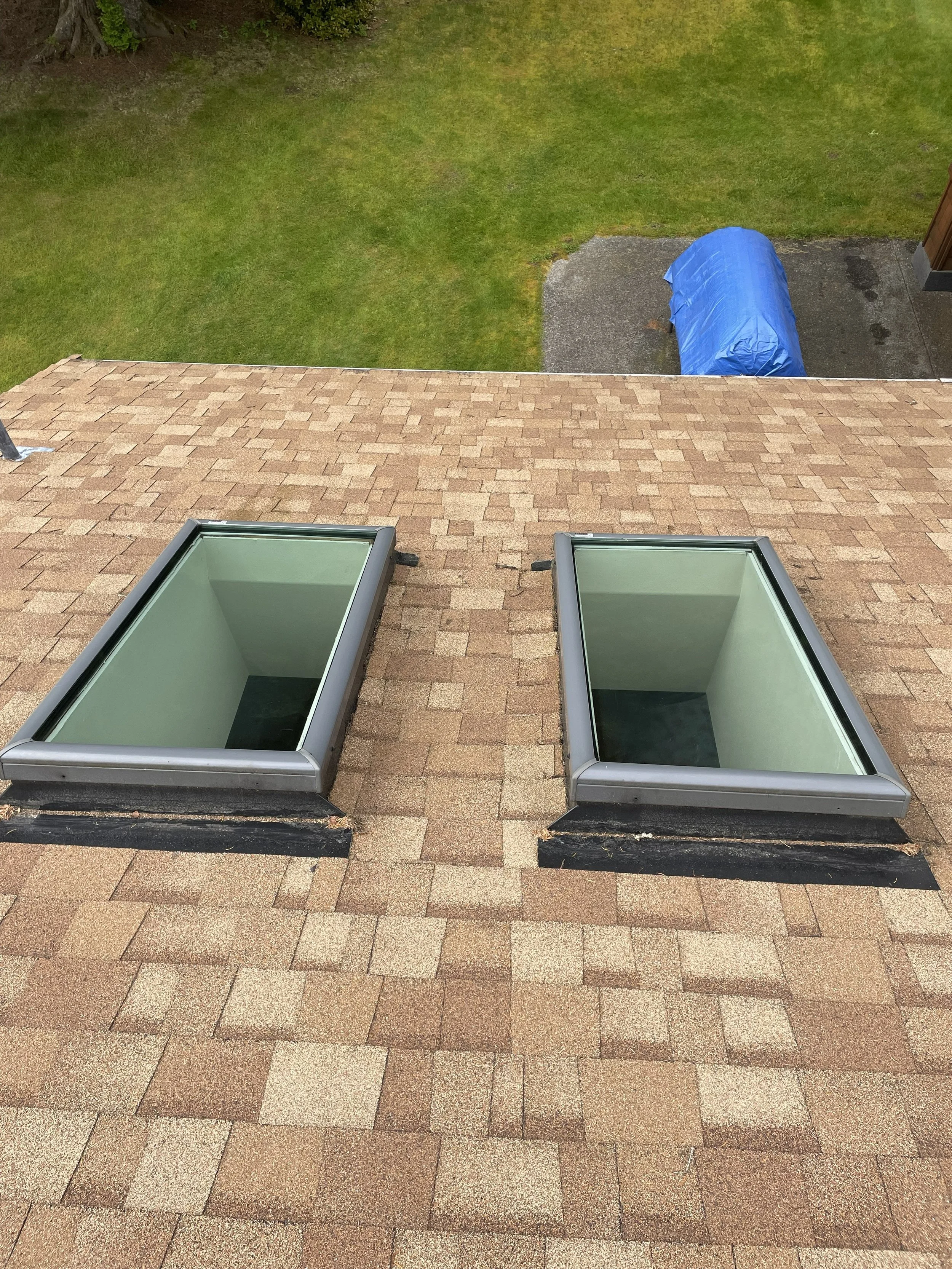 Two skylights on a shingled roof with a green yard and a blue tarp-covered object in the background.