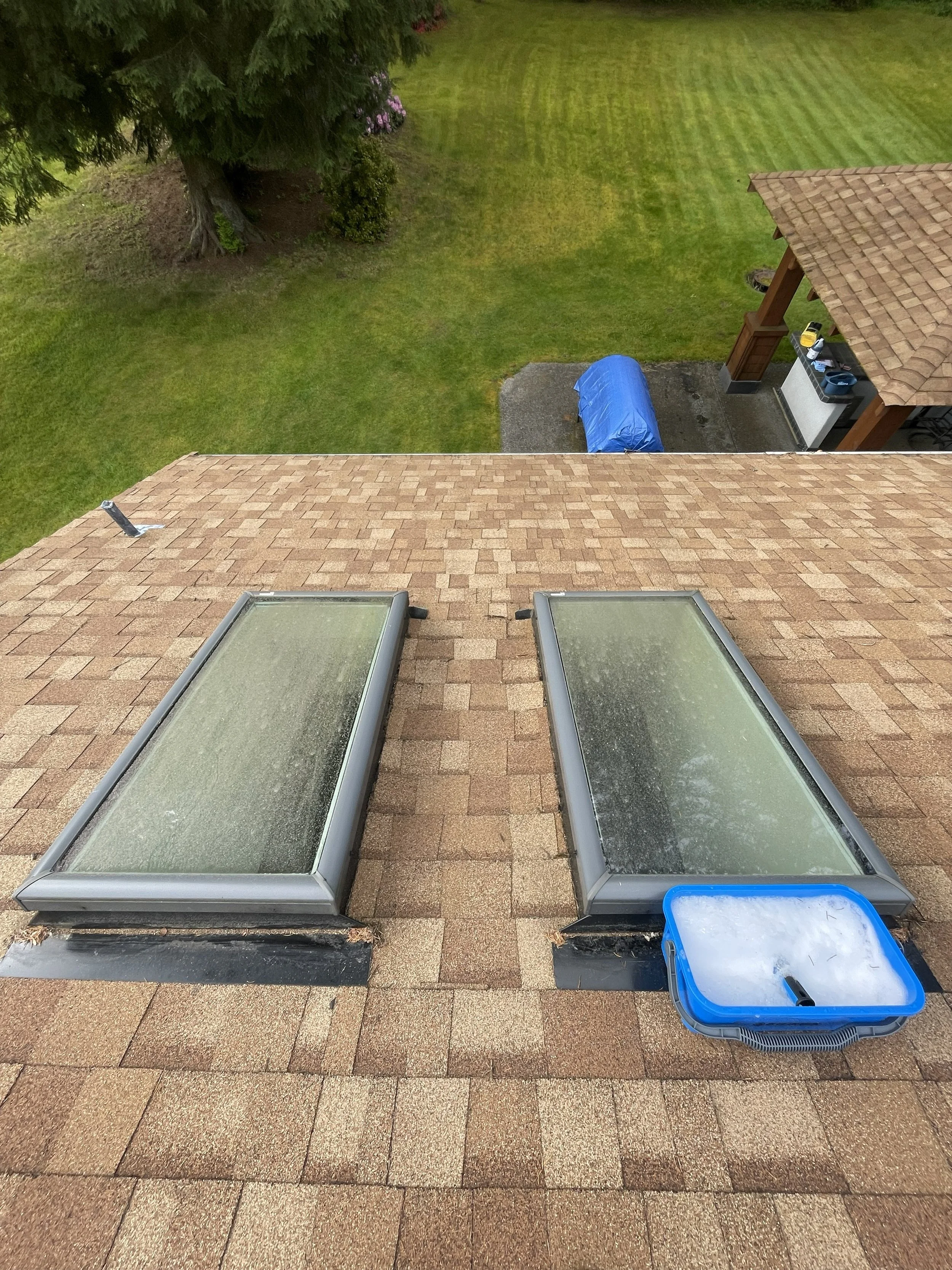 View from a rooftop showing two skylights, a plastic bin with snow, and a backyard with grass, a tree, and a covered object.