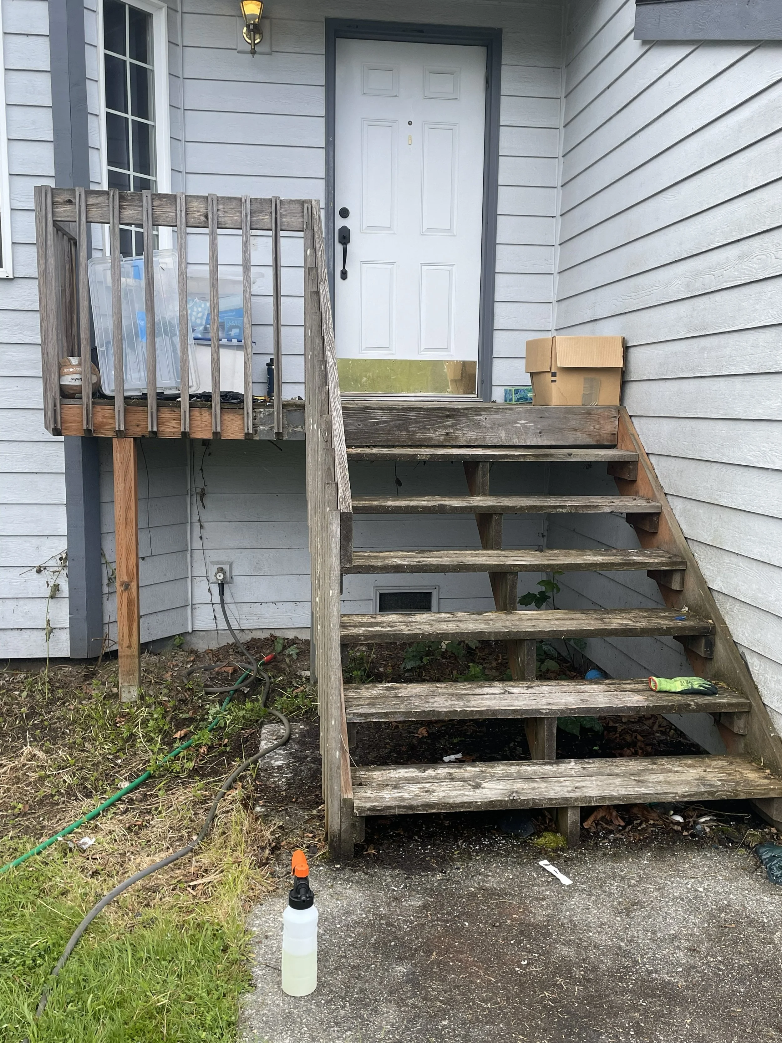 Wooden stairs leading to a white front door with a black handle and lock, attached to a house with gray horizontal siding, and a small porch area with some objects and a cardboard box on it.