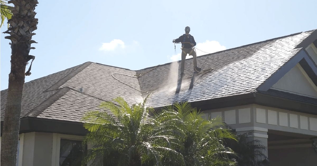 A person on a harness cleaning or inspecting a house roof with a high-pressure water hose.