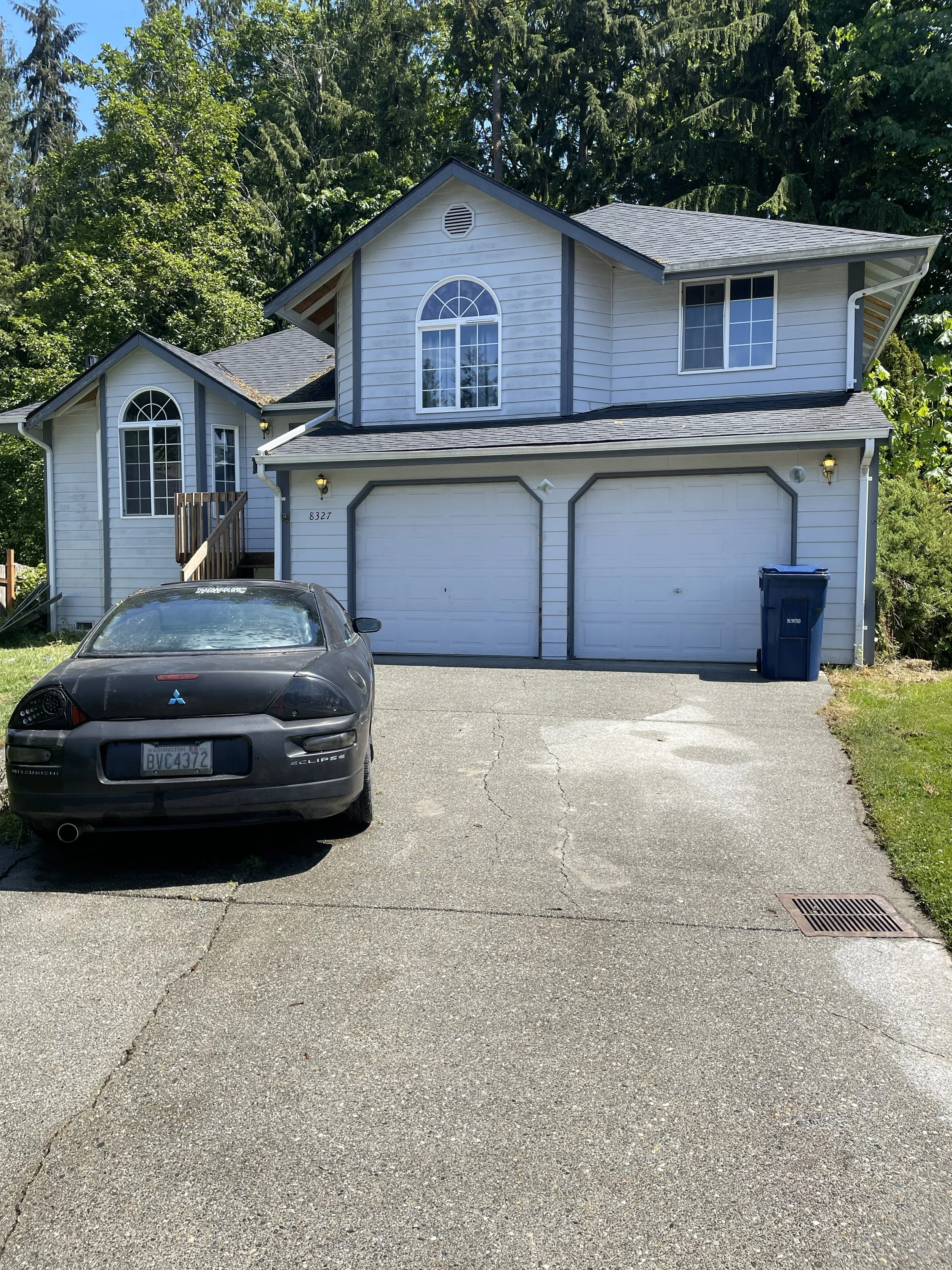 A two-story suburban house with white siding, two garage doors, and a black Mitsubishi Eclipse parked in the driveway. There is a blue trash bin next to the house, and the house number 8327 is visible. The background includes tall green trees.