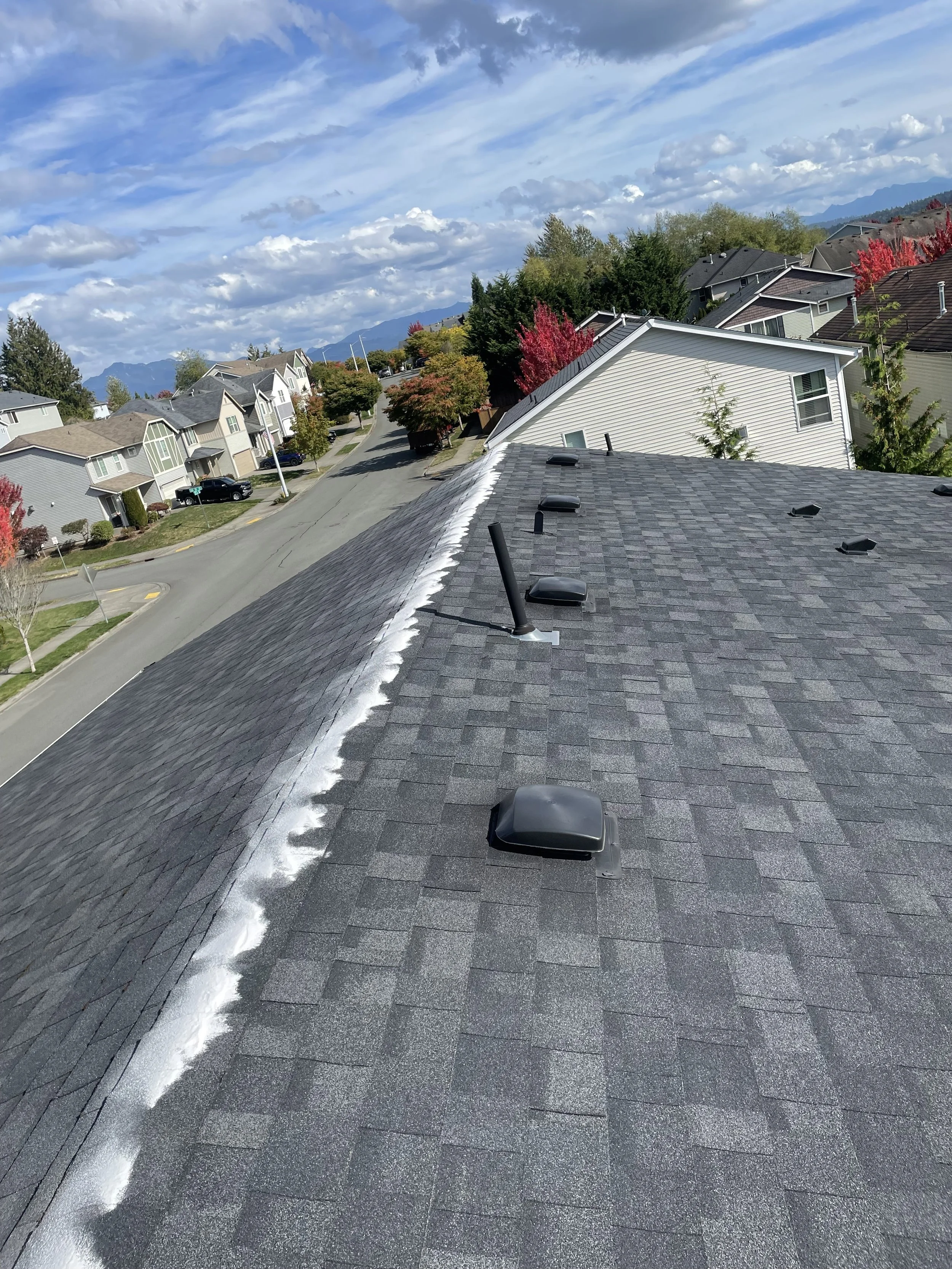 View from a rooftop showing asphalt shingles, vents, and pipes with a suburban neighborhood and mountain range in the background under a partly cloudy sky.
