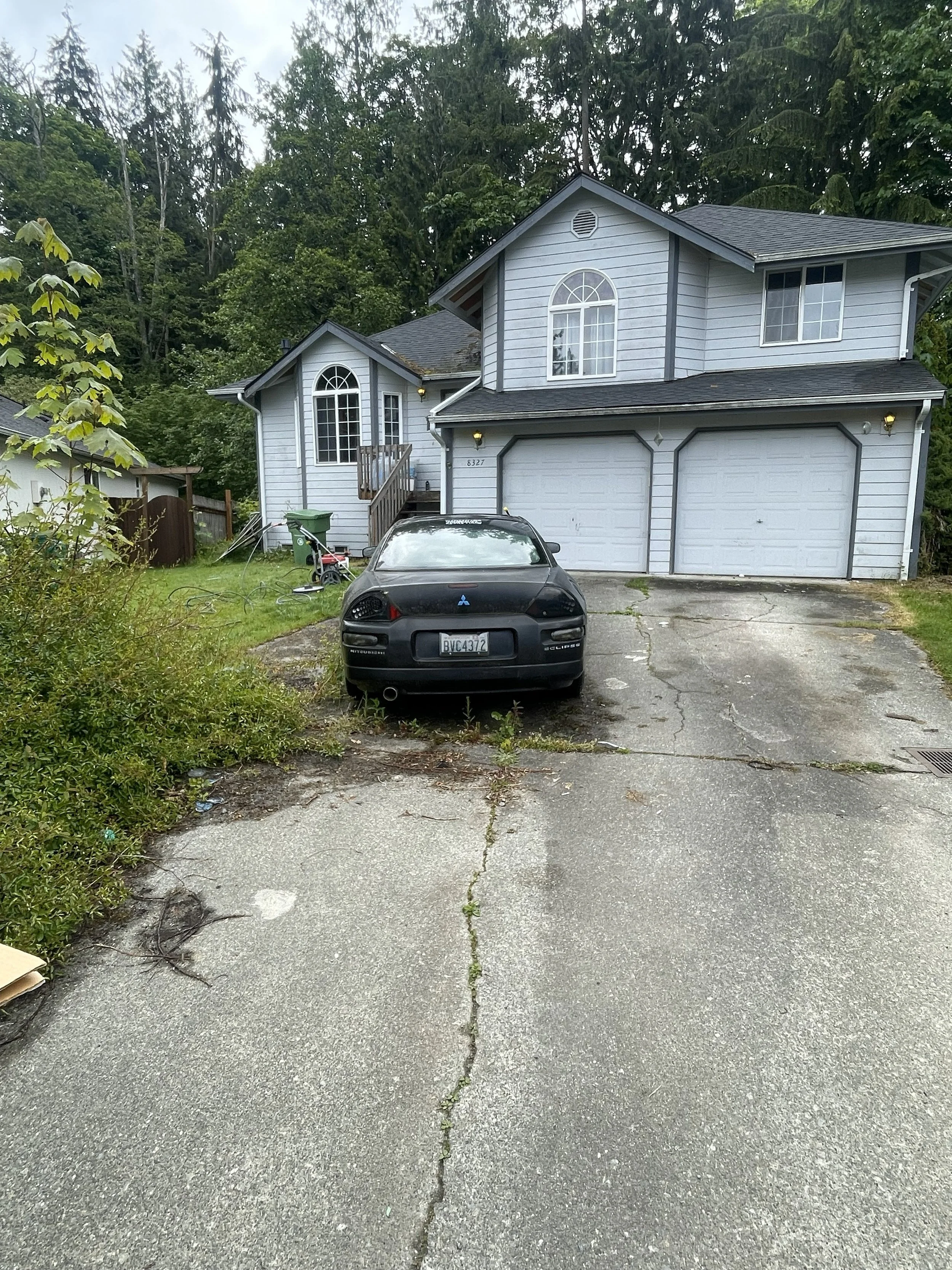 A two-story house with light blue siding and white garage doors. There is a black car parked in the driveway in front of the house. The driveway has visible cracks and weeds growing in it. The surrounding area has green trees and bushes.