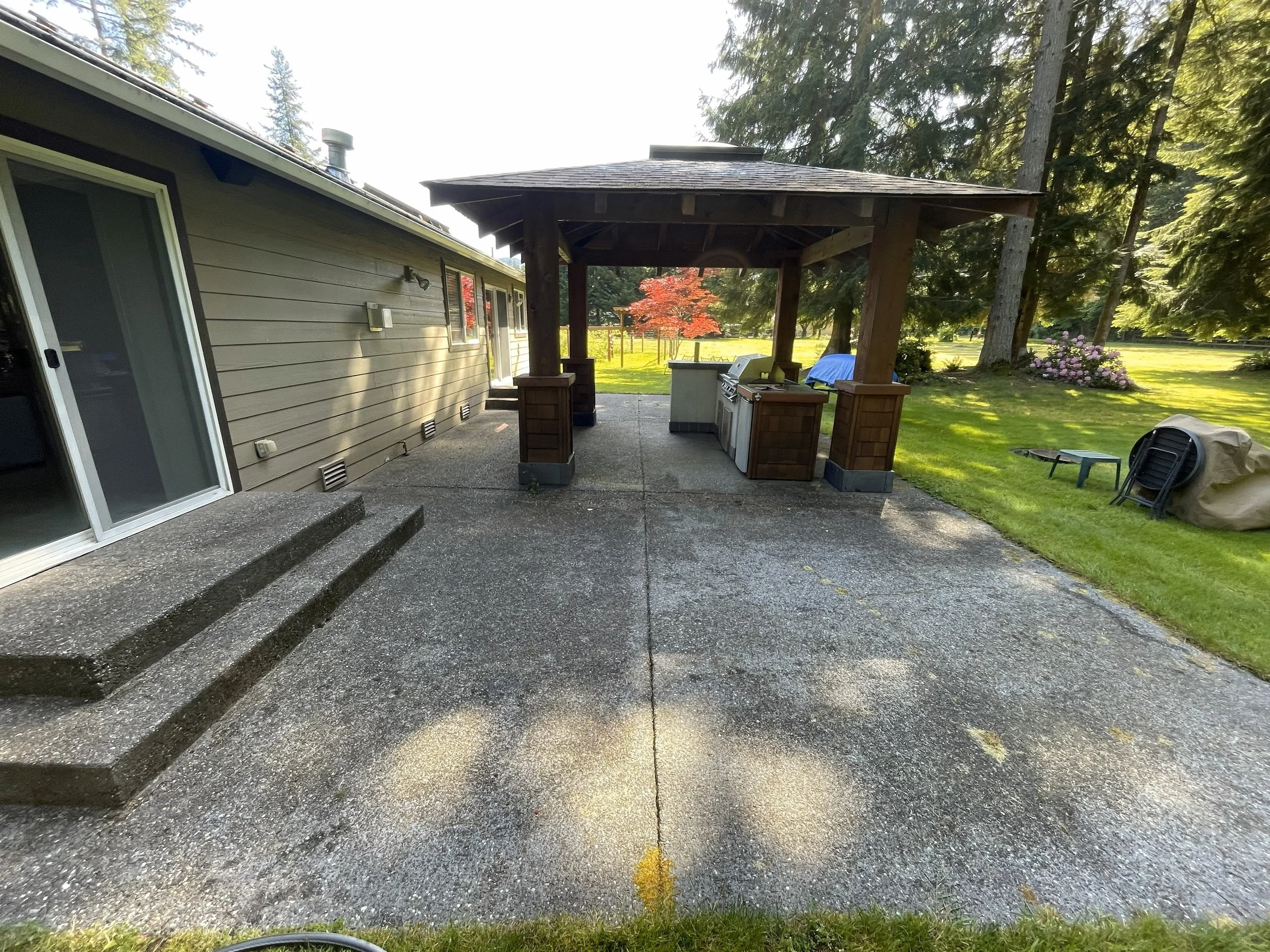 View of a backyard patio with a concrete surface, a covered outdoor kitchen area with a grill, countertop, and storage, adjacent to a house with sliding glass doors, and a grassy lawn with trees and pink flowers in the background.