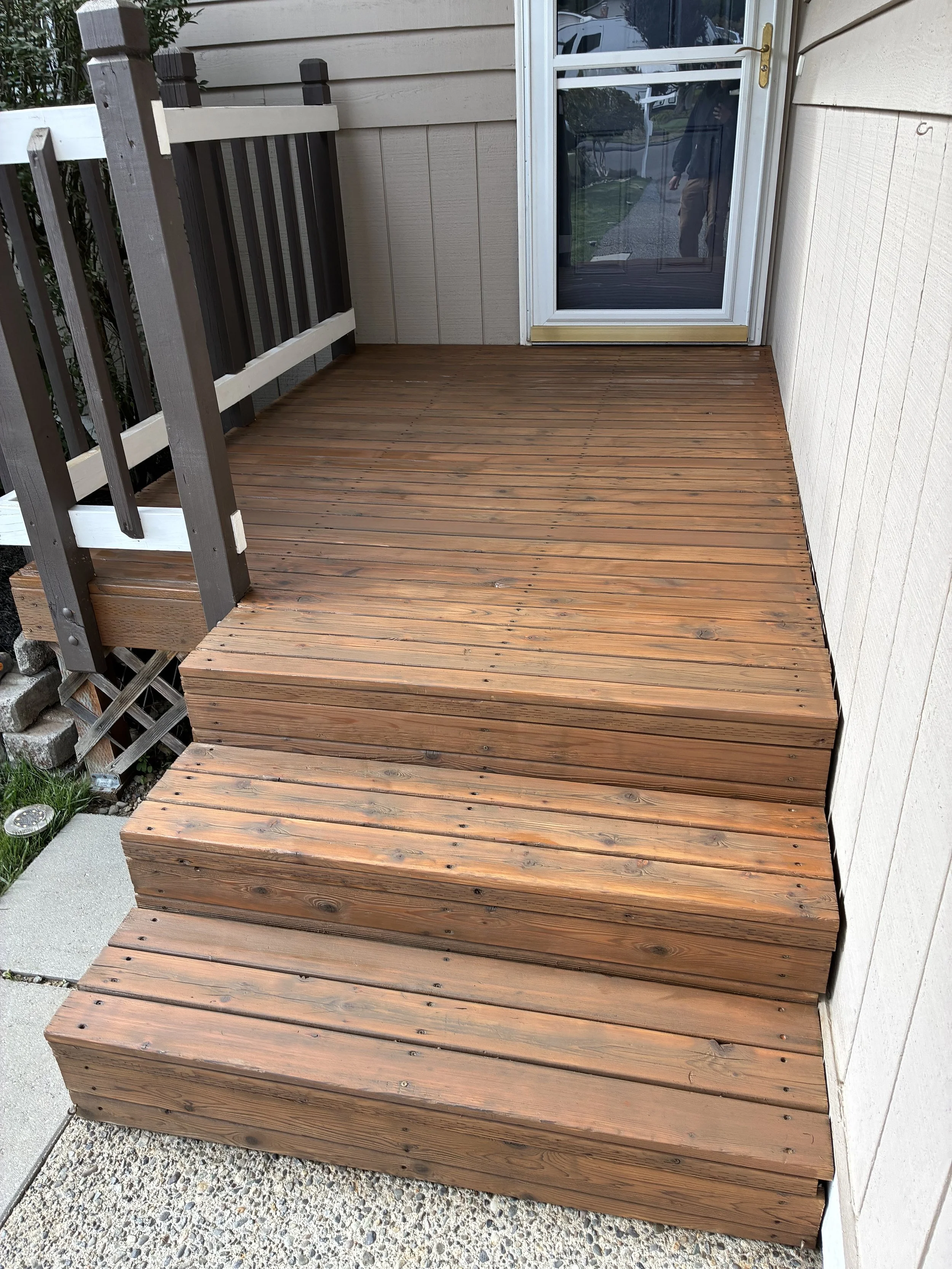 Three wooden steps leading up to a small balcony or deck area outside a house, with a glass door and a brown and white railing on one side.