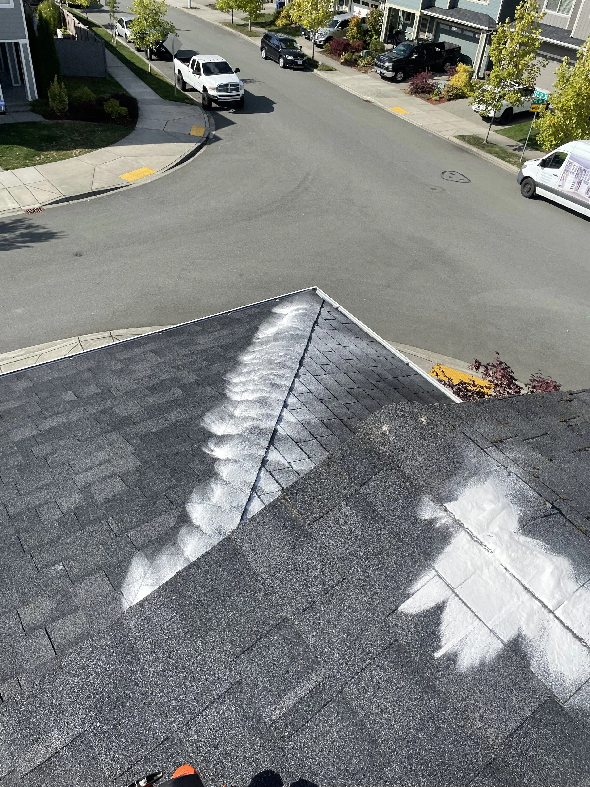View of a residential neighborhood street from a rooftop, showing houses, parked cars, and trees, with some roofing work visible.