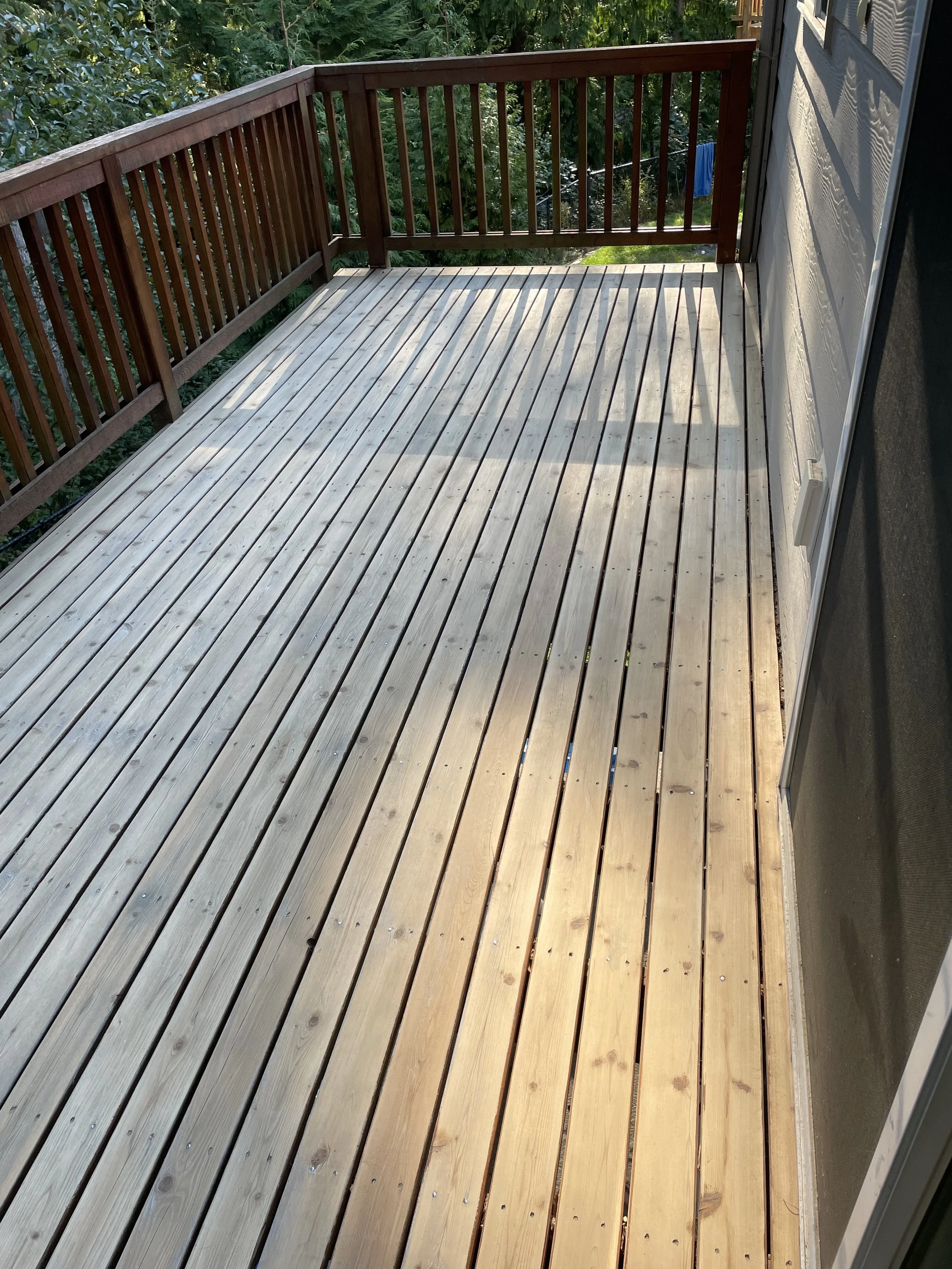 Newly installed wooden deck with railing and some greenery in the background.