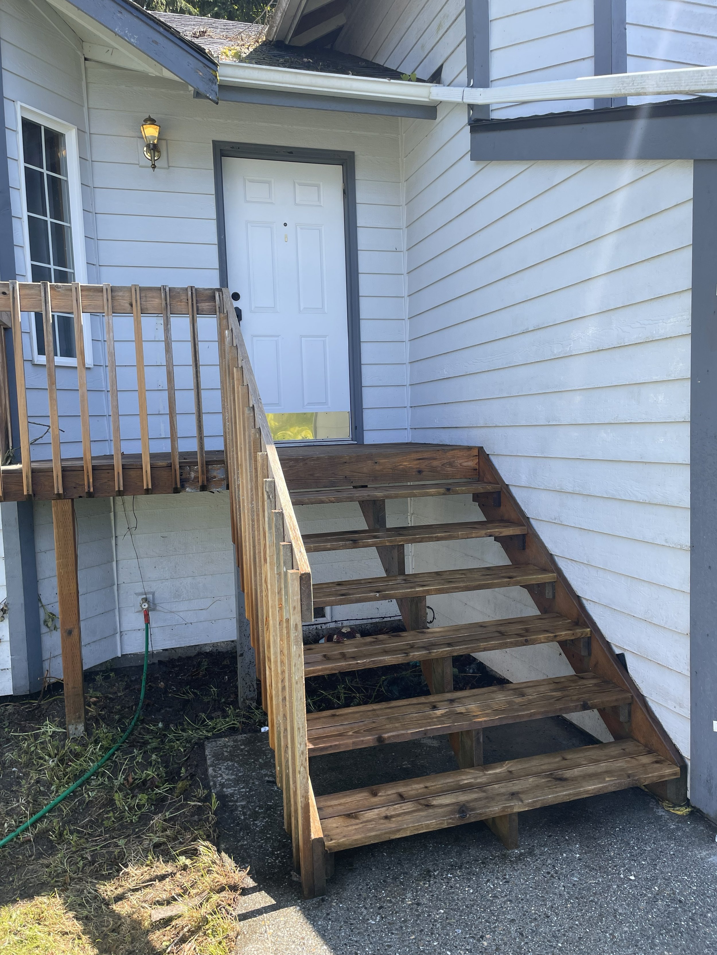 A small wooden staircase leading to a white back door with a small porch. The staircase has four steps, and there is a new railing on the left side. The house has white horizontal siding, and a lantern-style outdoor light is mounted near the door. The porch area is elevated, and there is a garden hose on the ground next to the house.