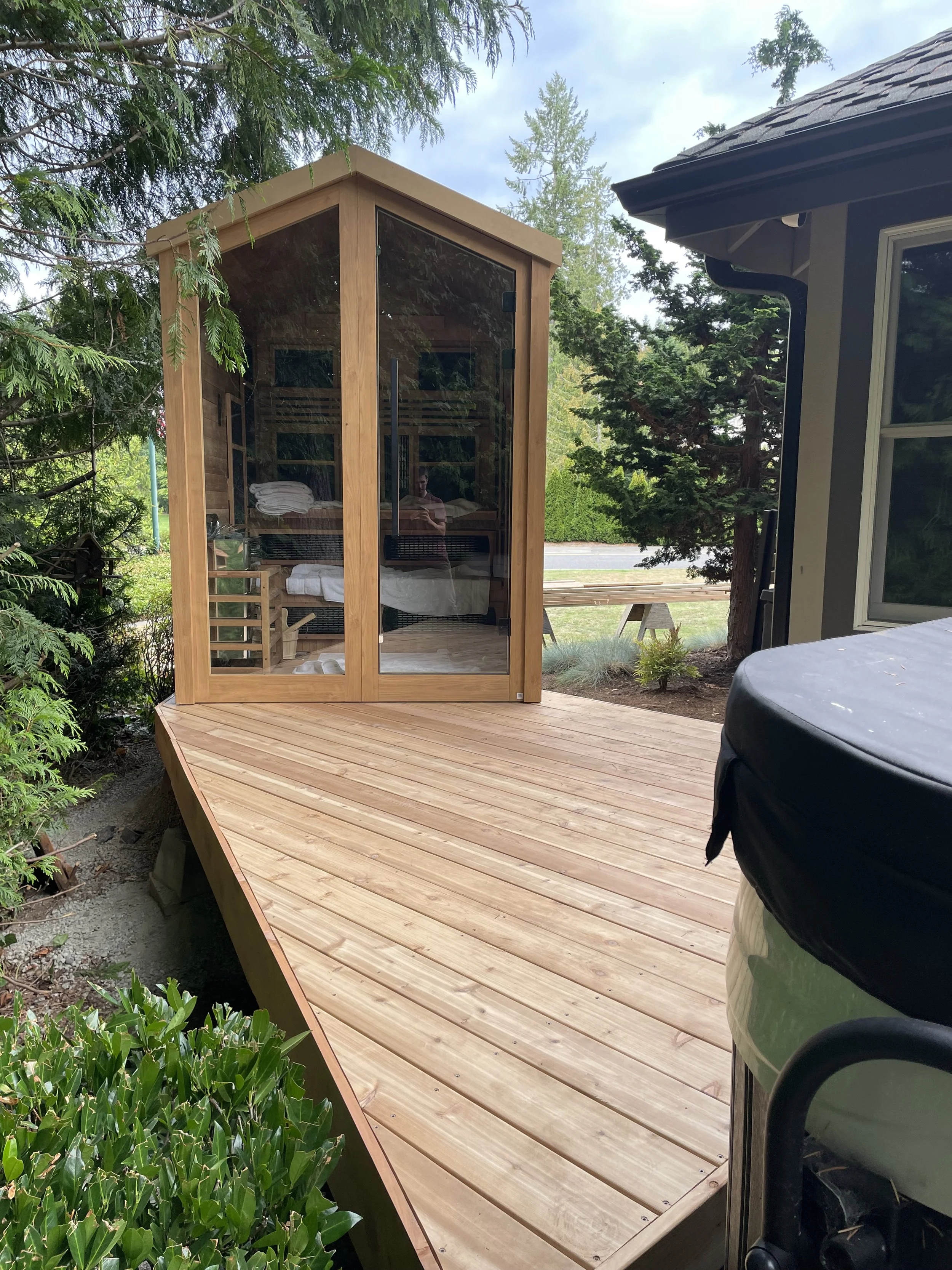 A wooden outdoor deck with a glass-enclosed sauna located in a backyard. The sauna has a natural wood finish, and inside, towels are visible. Surrounding greenery and a neighboring house are also visible.