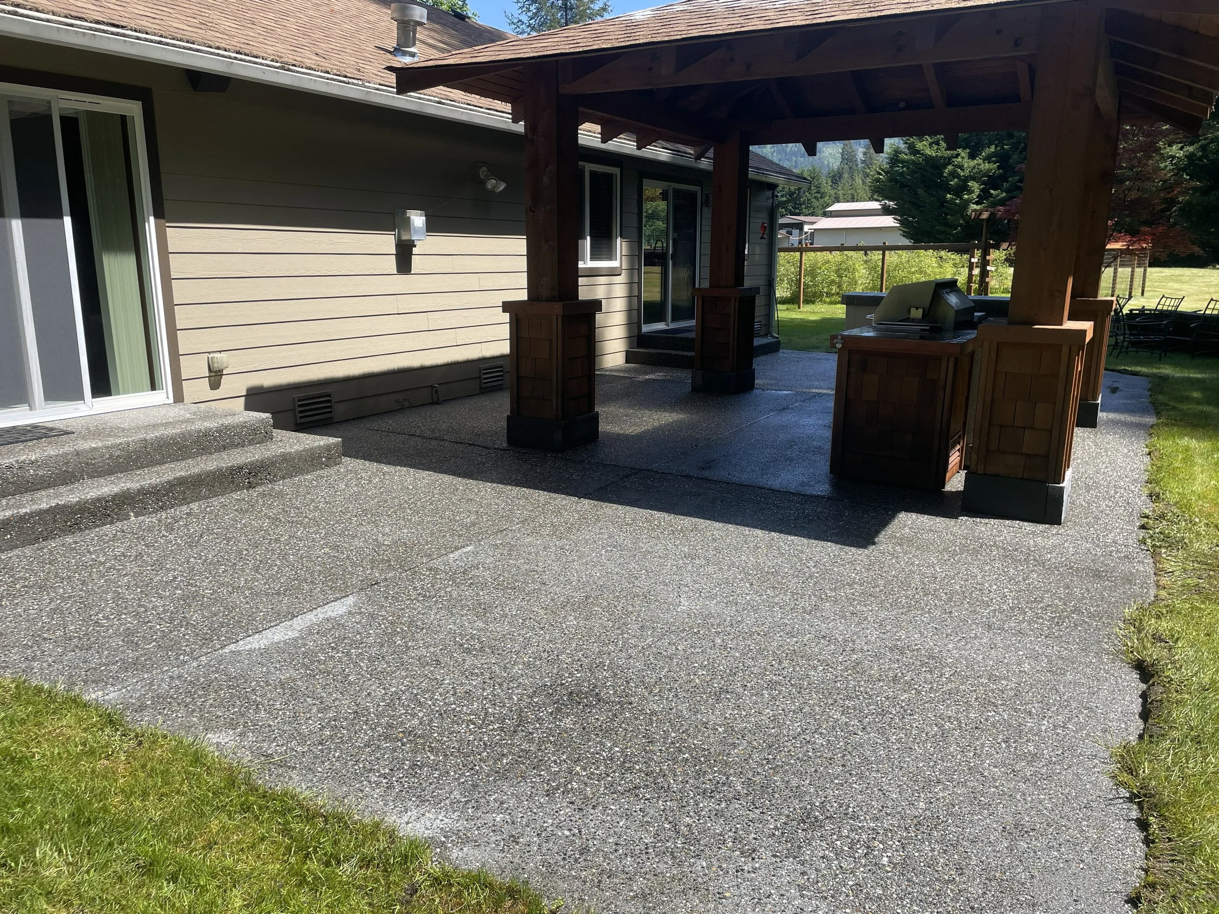 Backyard patio with concrete flooring, wooden covered outdoor kitchen area, and sliding glass door leading inside house; grassy yard with trees in the background.