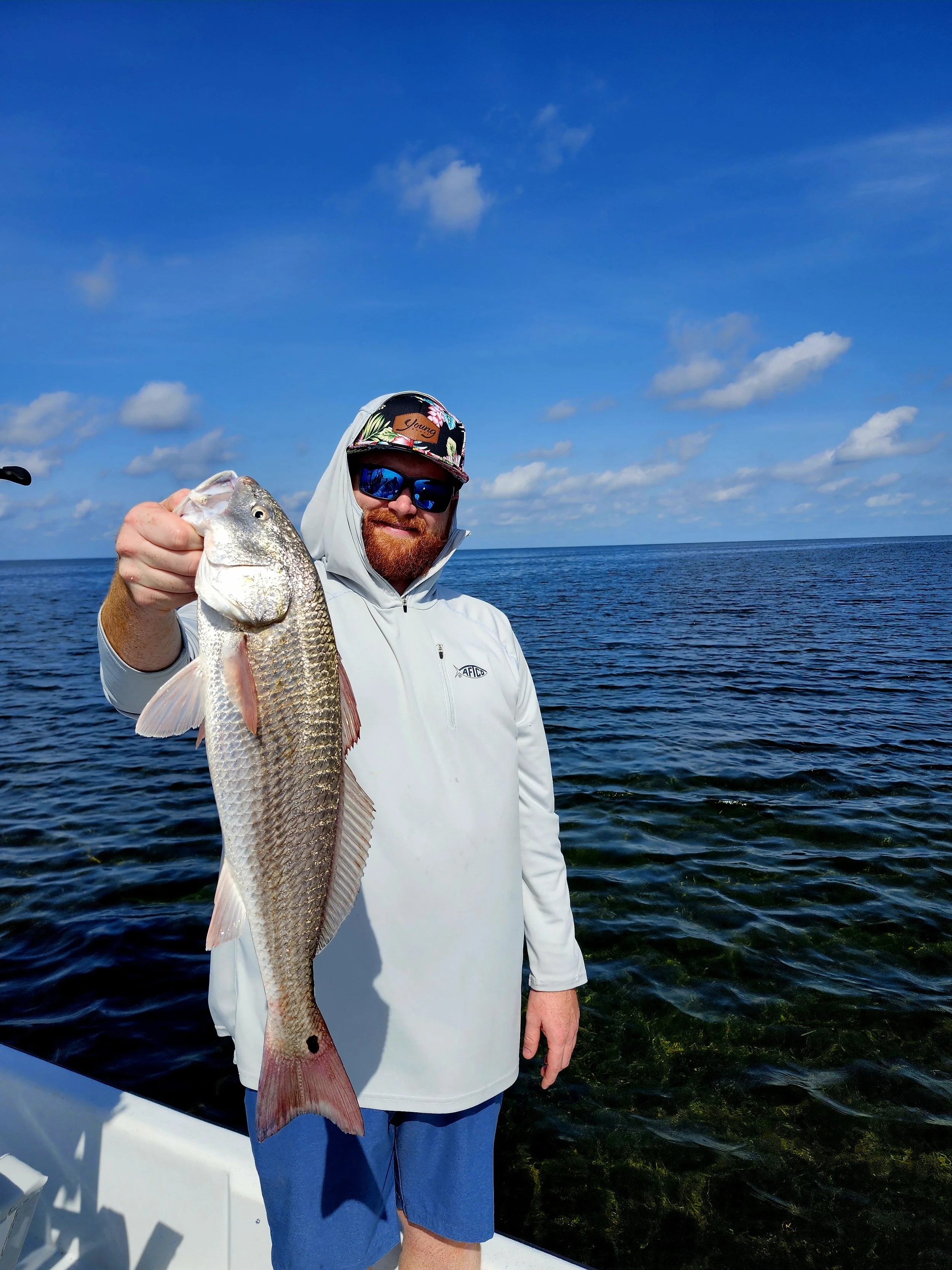 A man with a beard wearing sunglasses, a gray hoodie, and a colorful hat, holding a large fish on a boat in the ocean under a blue sky with clouds.