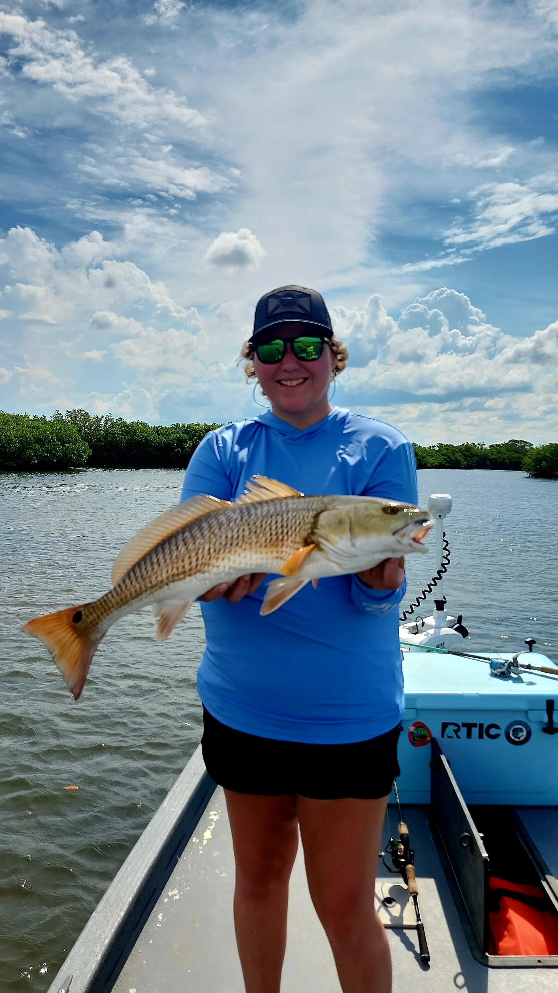 Person holding a large fish on a boat in a waterway with trees and cloudy sky in the background.
