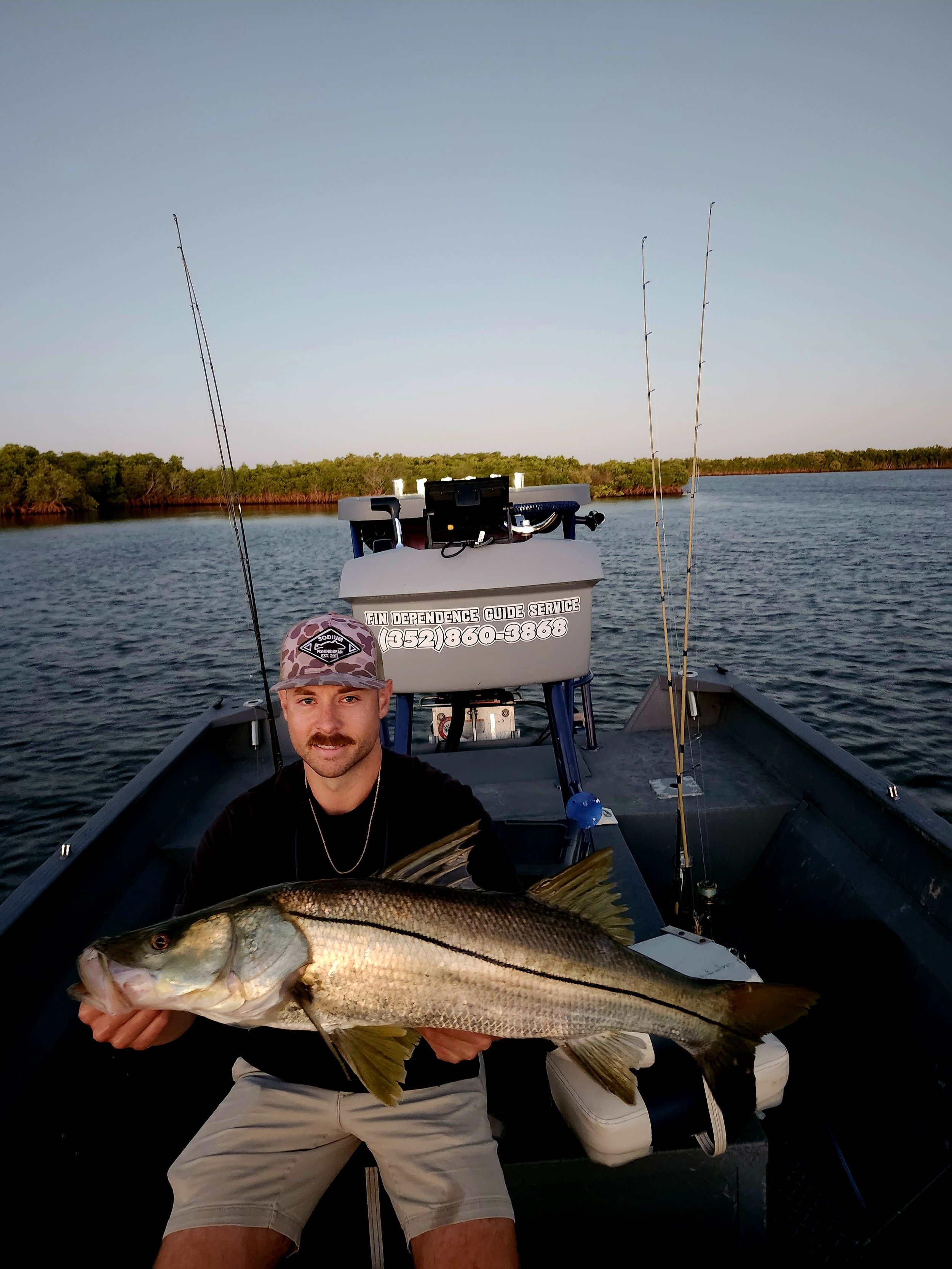 A man on a boat holding a large fish he caught, with fishing rods visible in the background and water and trees in the distance.