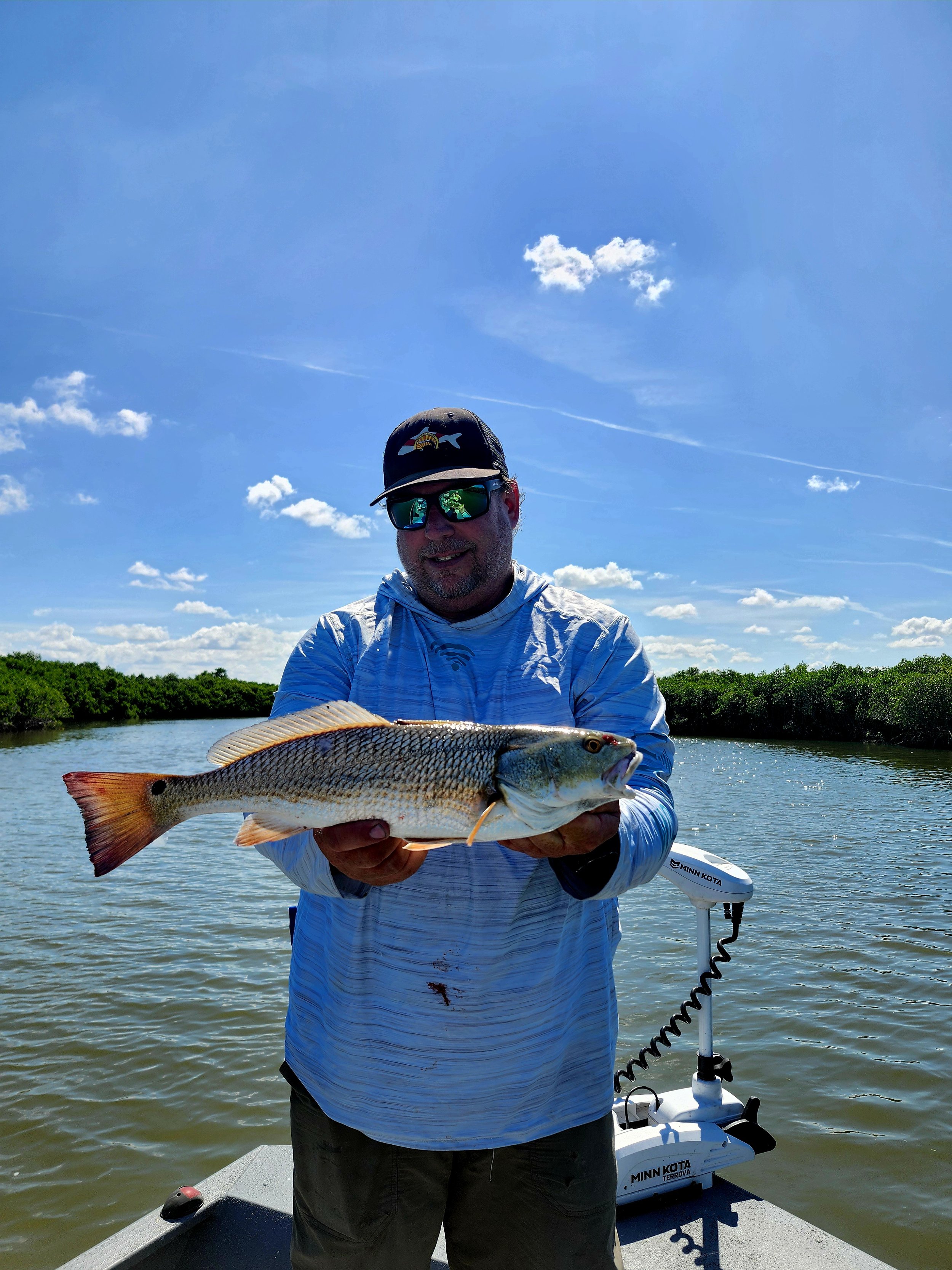 A man wearing sunglasses, a cap, and a light blue shirt stands on a boat in a river holding a large fish with both hands. The background features a clear blue sky with some clouds and green trees on the riverbanks.