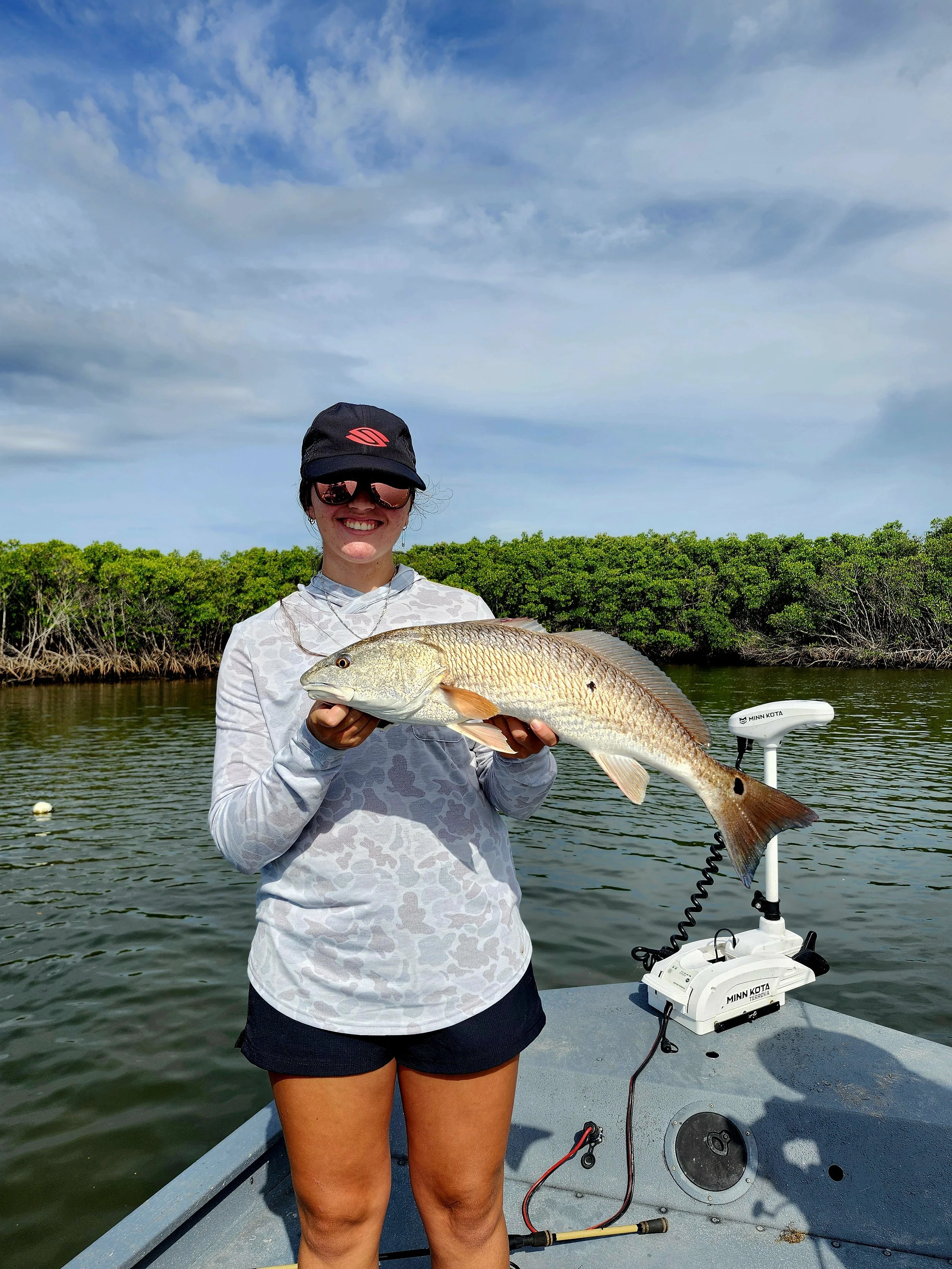 A woman on a boat holding a large fish she caught, with water and green trees in the background.