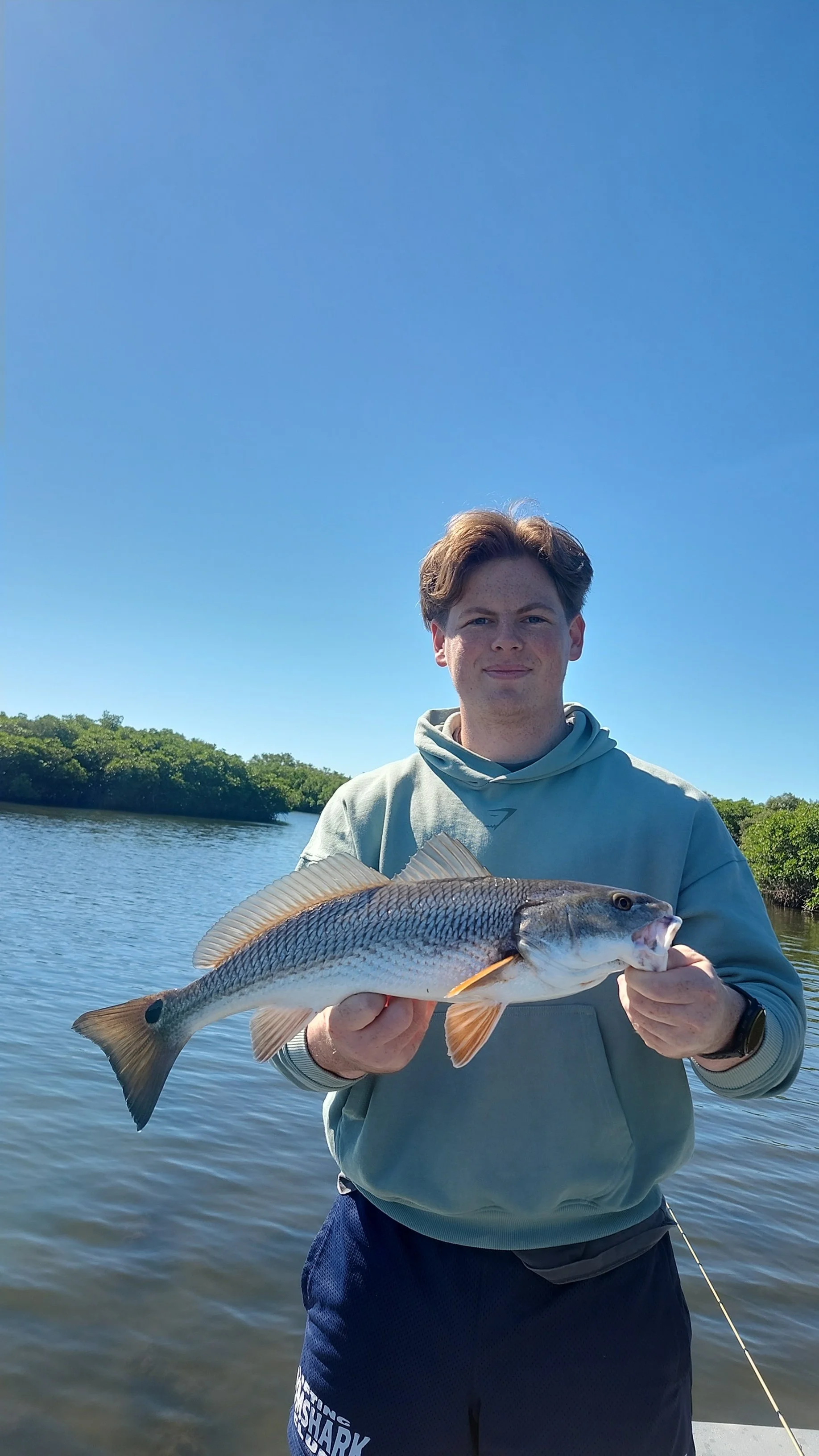 A young man on a boat holding a large fish with a body of water and green trees in the background under a clear blue sky.