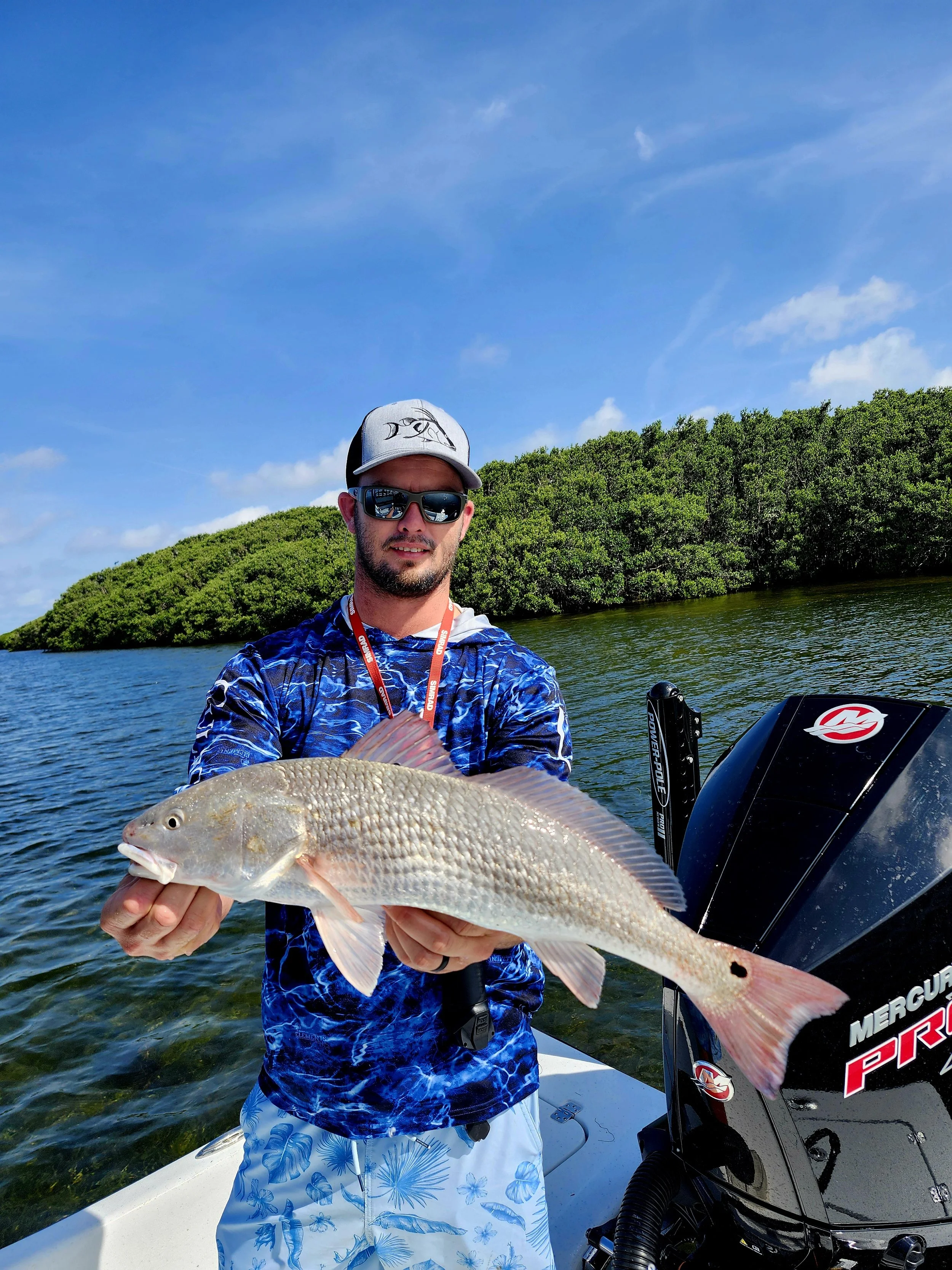 Man in sunglasses holding a large fish on a boat, with water and green bushes in the background under a blue sky.