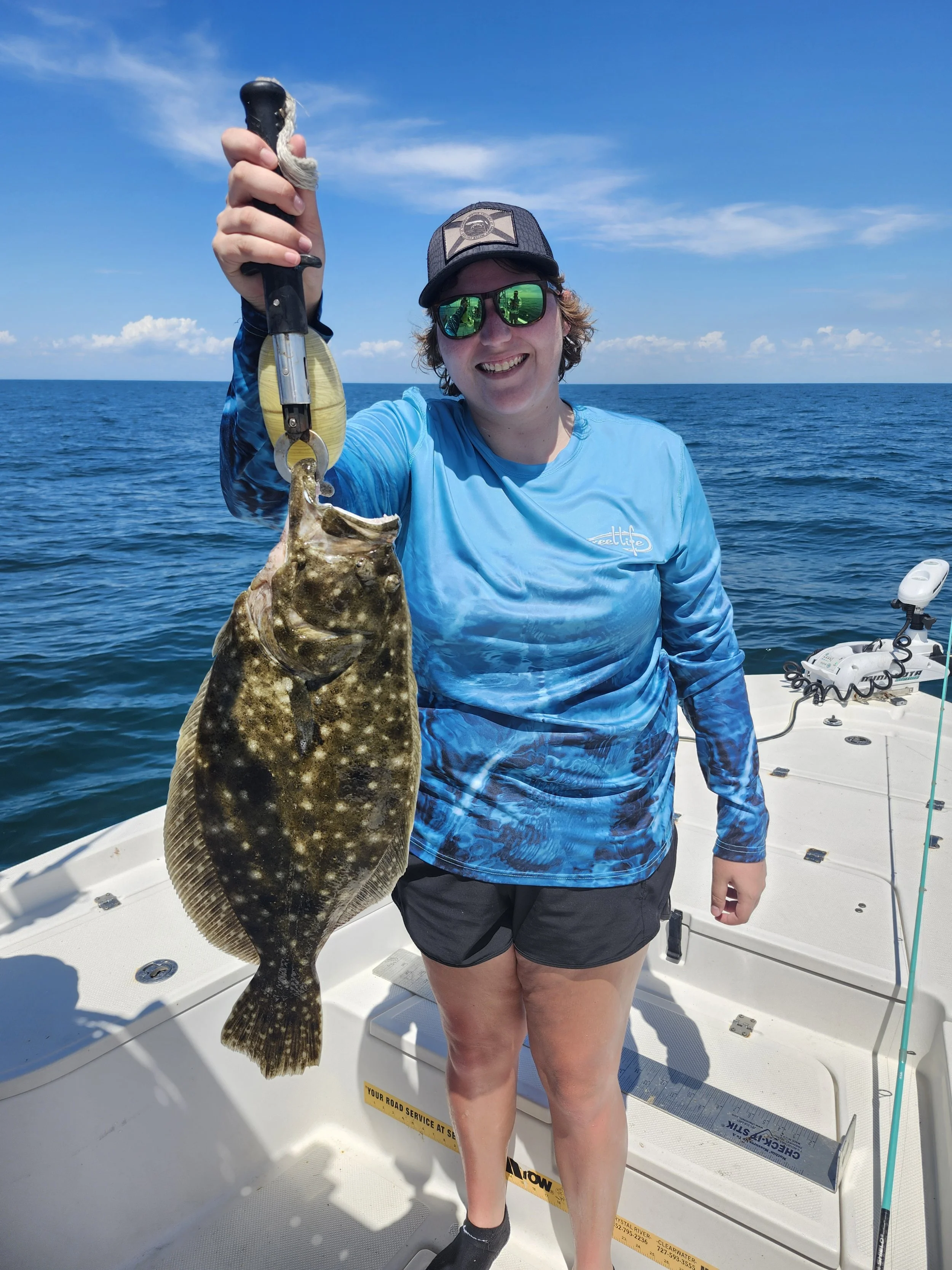 A woman holding a fish on a boat during a sunny day, smiling and wearing sunglasses, a blue shirt, and a cap with a background of the ocean and blue sky.