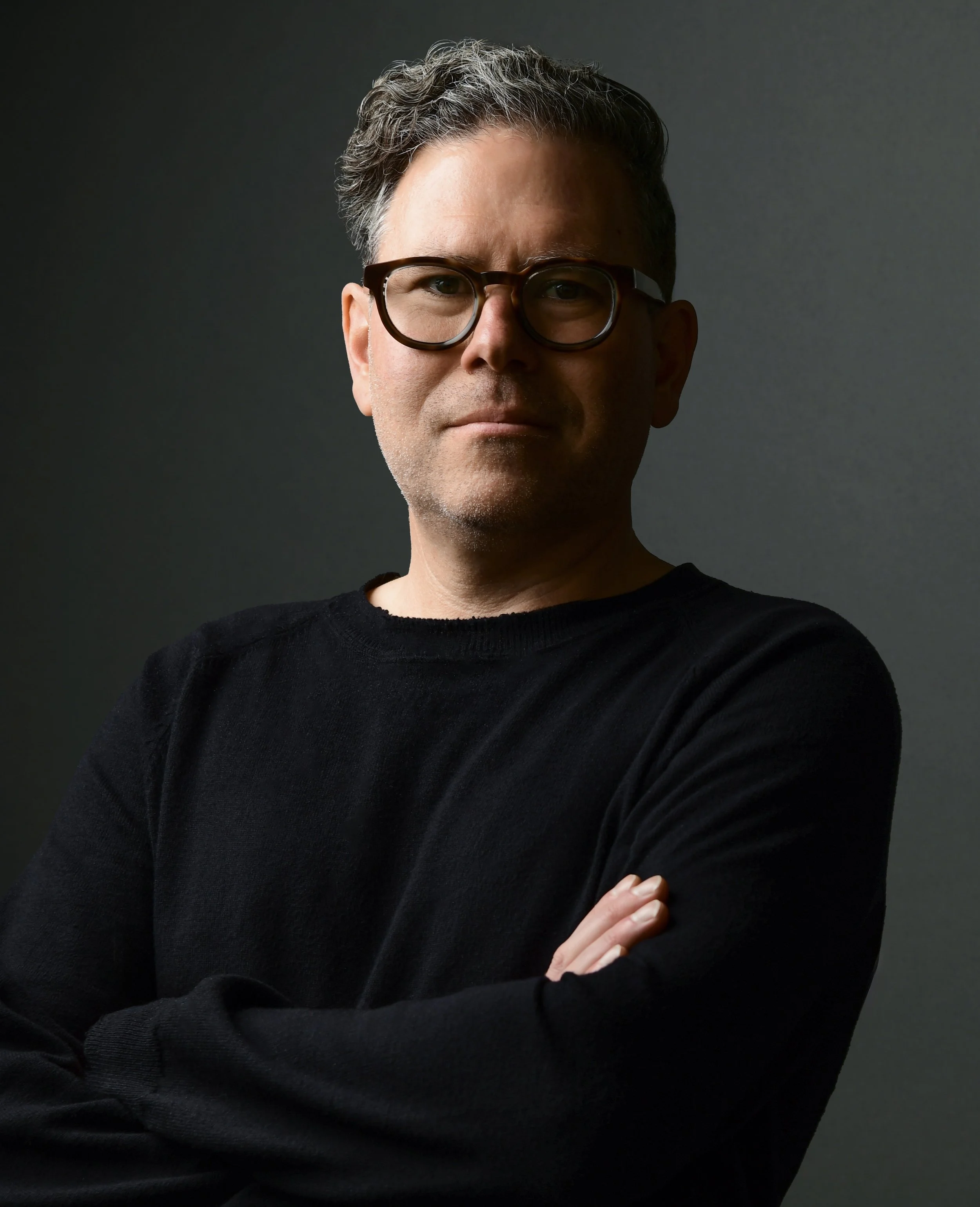 A man with glasses and dark hair wearing a black sweater, standing with arms crossed against a neutral background.