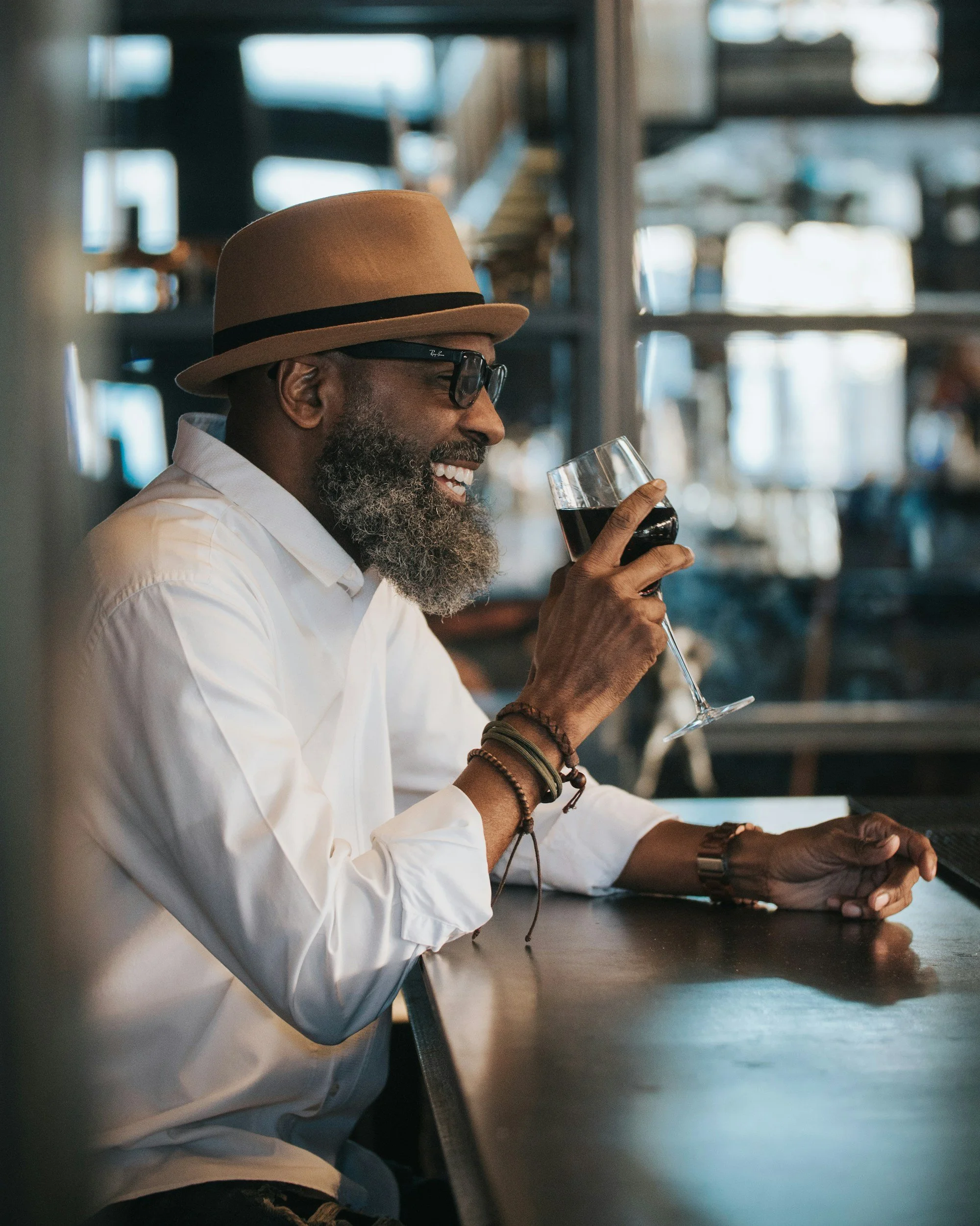 A man with a gray beard, sunglasses, and wearing a tan hat and white shirt is sitting at a bar or cafe, smiling and holding a glass of red wine.
