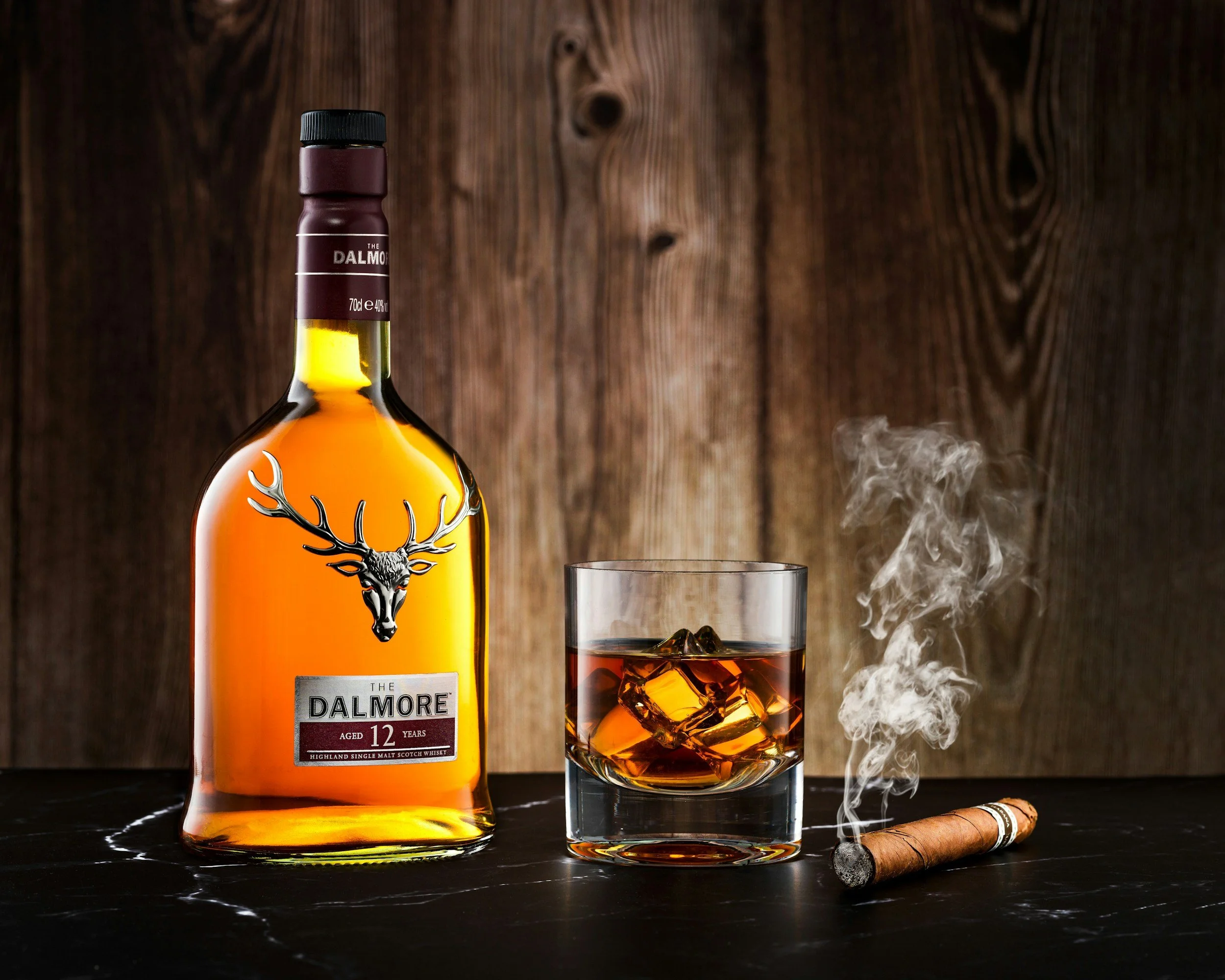 A bottle of Dalmore 12-year-old single malt Scotch whisky, a glass of whisky with ice, and a lit cigar emitting smoke, all placed on a black surface with a wooden background.