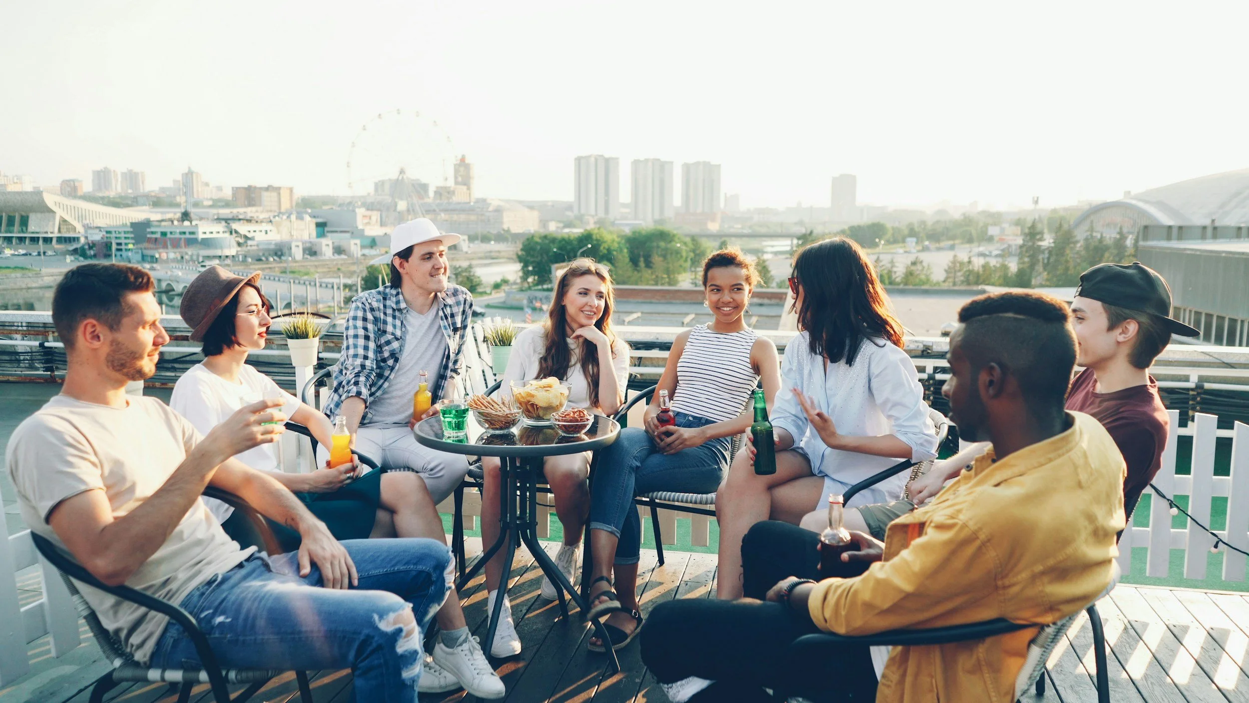 Group of friends sitting together on a rooftop deck, enjoying drinks and snacks, with a city skyline and Ferris wheel in the background.
