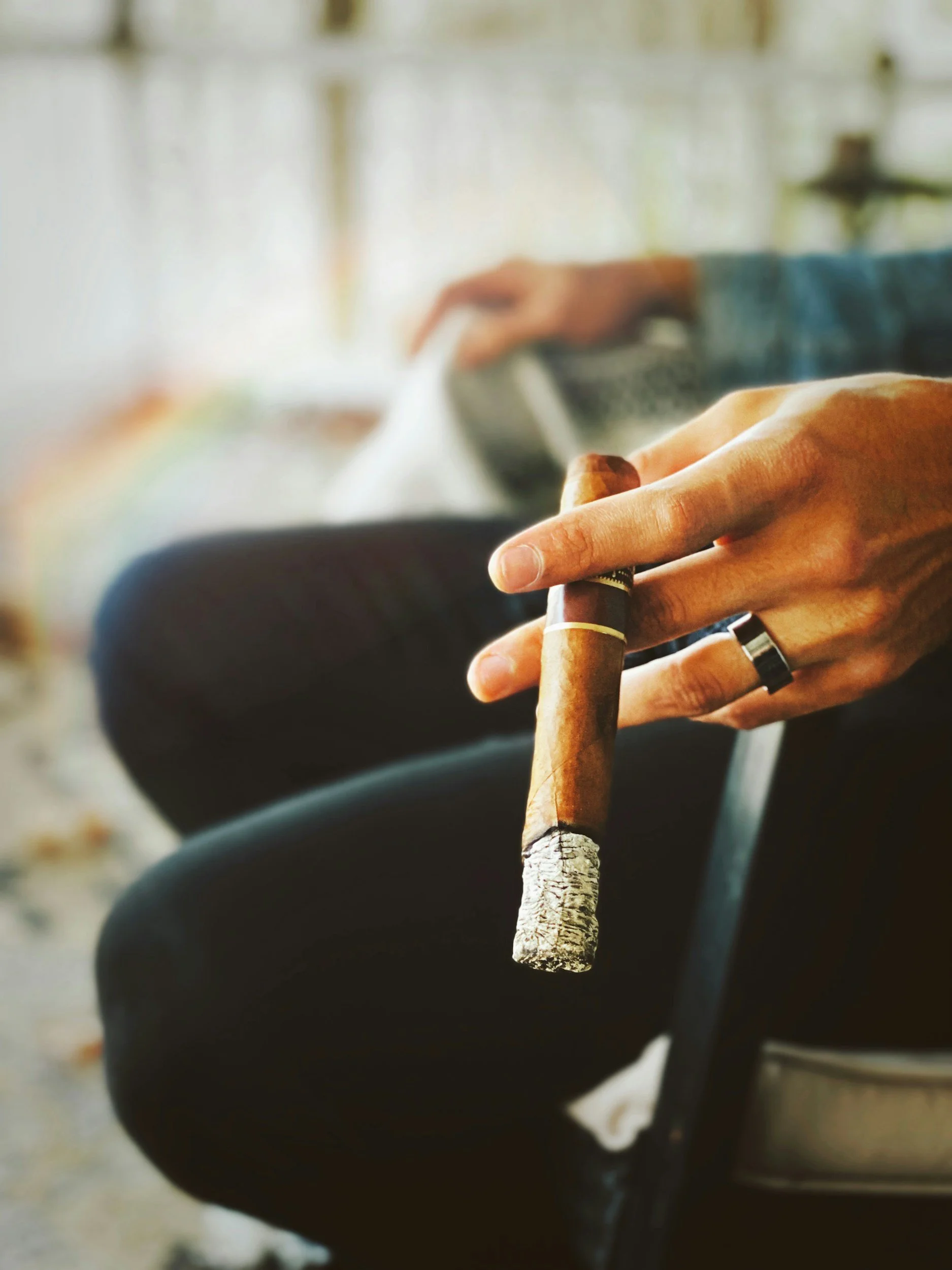 Close-up of a person's hand holding a cigar, with a blurred background of a person sitting and reading a book.