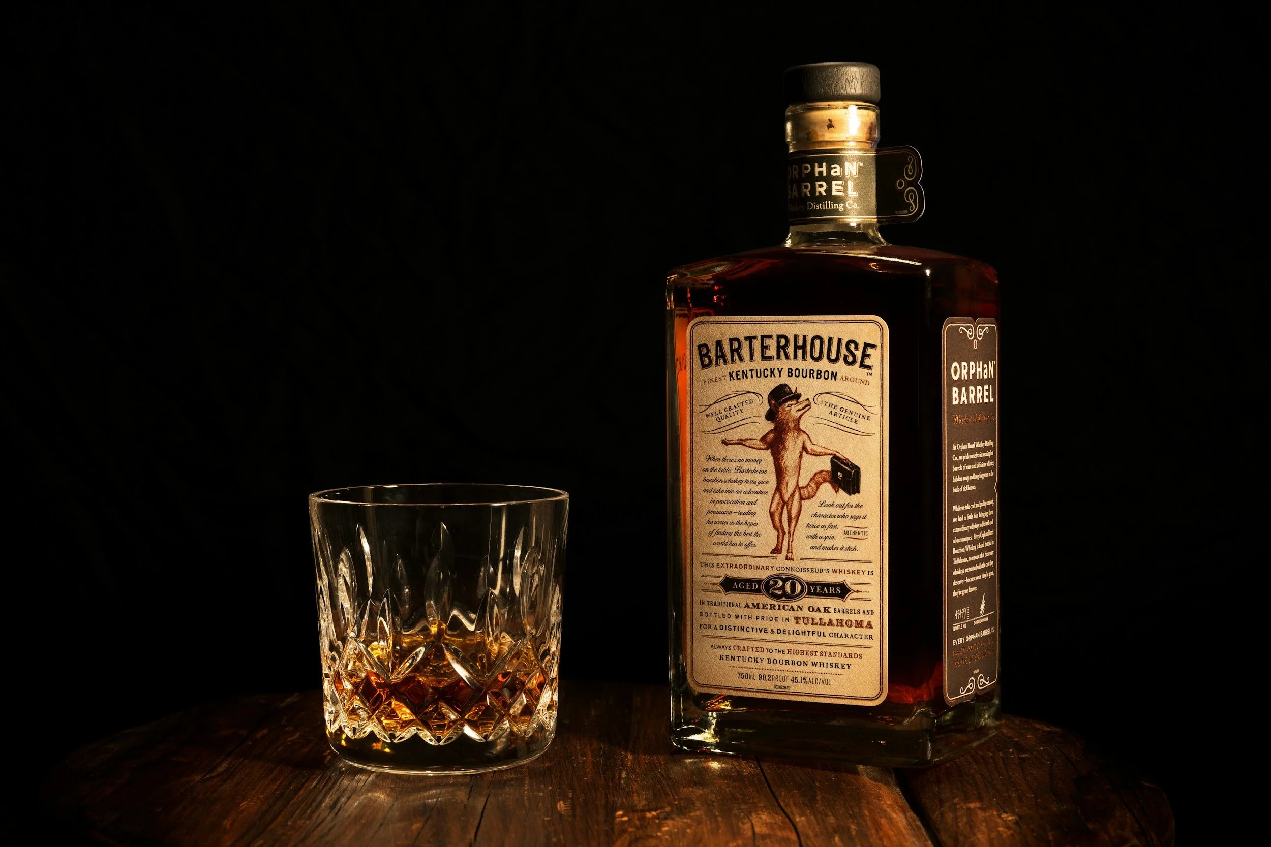 A bottle of Barterhouse Kentucky bourbon whiskey and a glass filled with bourbon on a wooden surface, with a dark background.