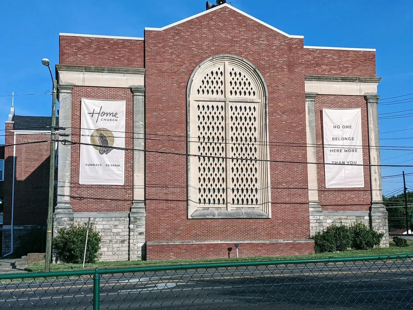 Red brick church building with large window in the center, flanked by two banners. The left banner reads 'Home Church Sundays 10:30AM', and the right banner reads 'No one belongs here more than you'.