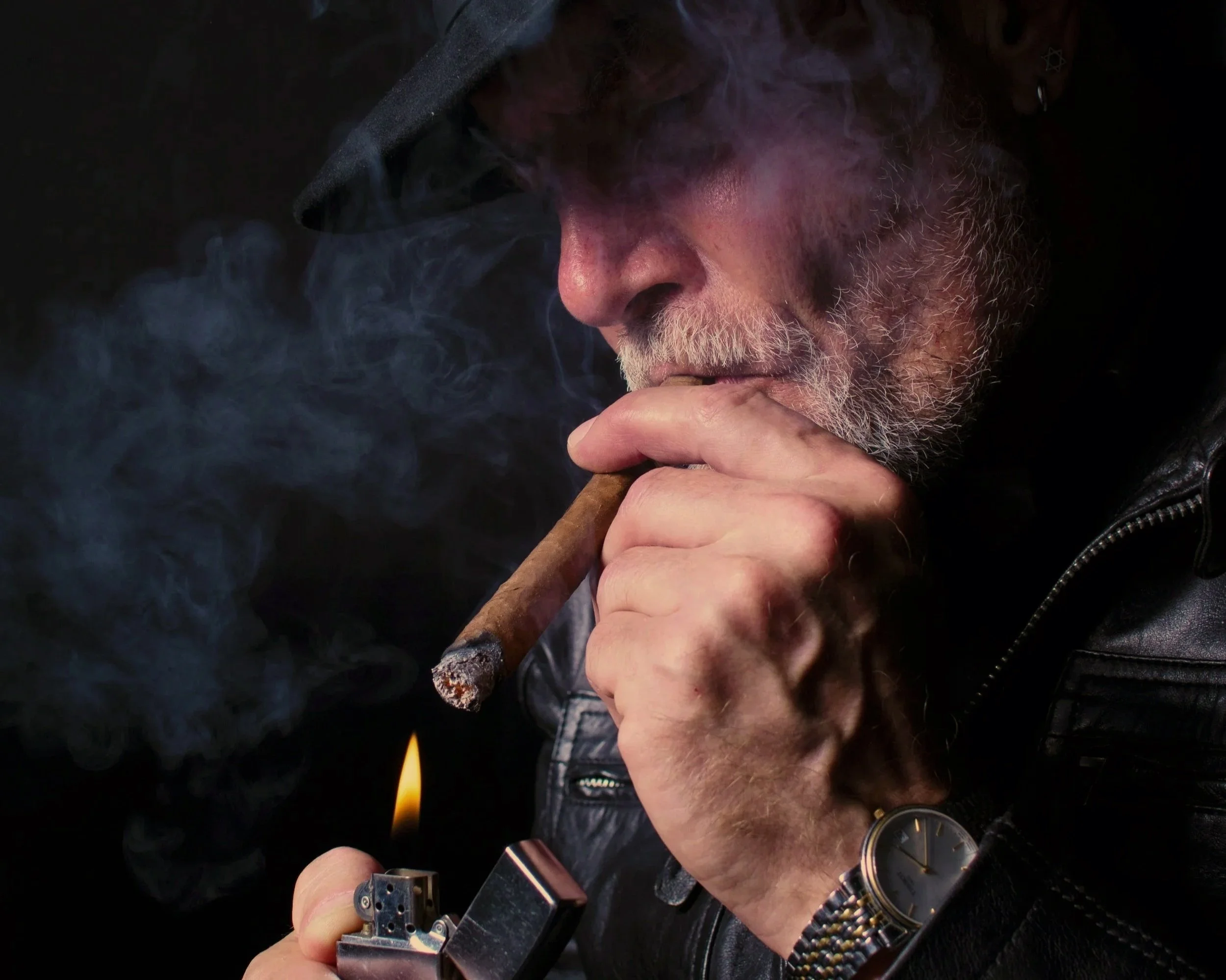 A man with gray hair, beard, and wearing a black leather jacket, black cap, and wristwatch, is lighting a cigar with a lighter. Smoke is visible around him against a dark background.